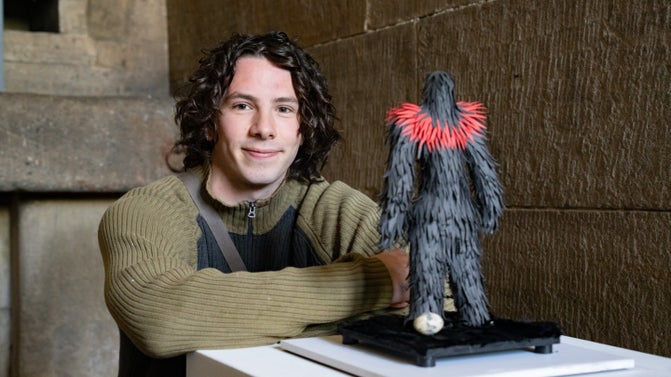 A young white man with a curly brown bob sitting behind a white plinth. On top of the plinth is a model of bird costume with black paper feathers and red paper feathers around the neck.