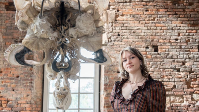 In the centre is a giant wig made from natural fibres which is suspending from the ceiling. Behind is exposed brickwork and to the right of the wig is a woman in a brown and blue striped shirt looking into the camera.