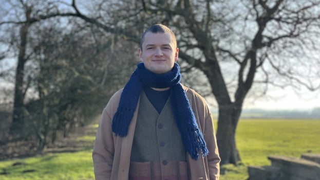 A white male with very short brown hair in a camel coat, olive green waistcoat and navy blue knitted scarf stands on grass with a tree in winter in the background