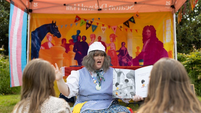 A woman in a white cap and blue and white checked smock sits in front of an open gazebo. She is holding a book and reading animatedly to children who are facing her