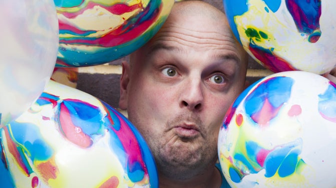A close up of a bald white man pulling a funny face, surrounded by multicoloured balloons.