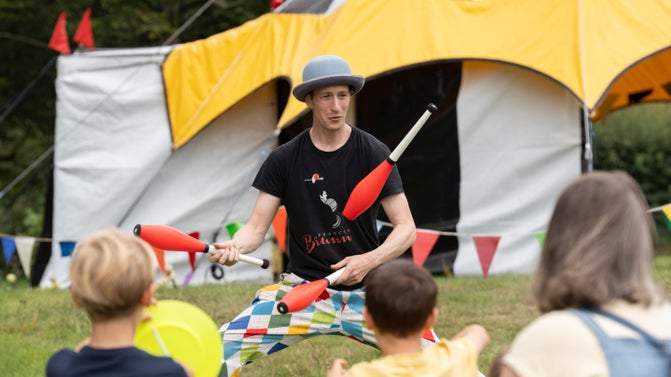 A man in a black tshirt and multicoloured trousers is standing in front of a black and white tent juggling three clubs