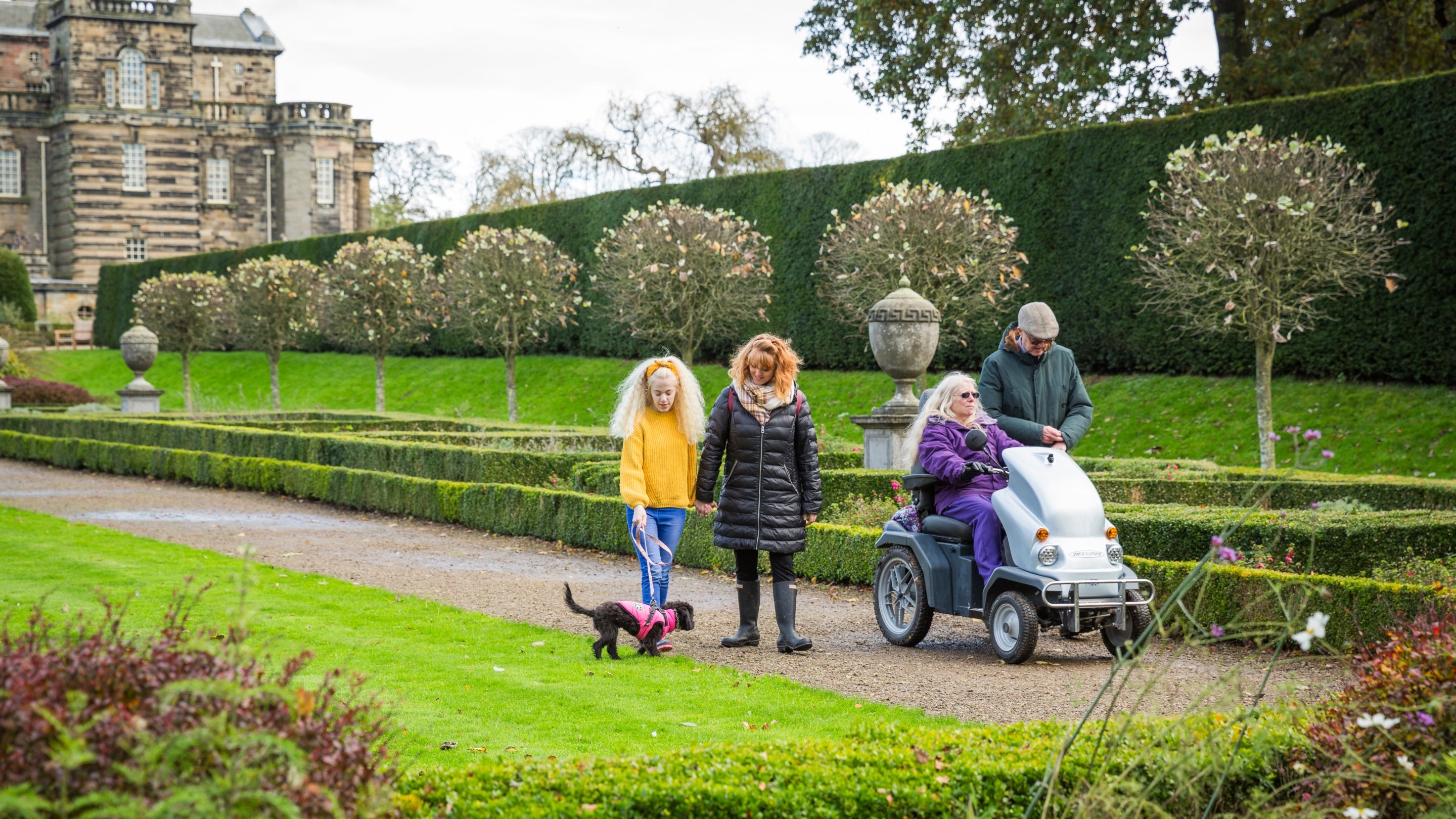 Four visitors walking down a garden path at Seaton Delaval Hall on an autumnal day, one person is in a mobility scooter and a young girls has a dog on a lead.