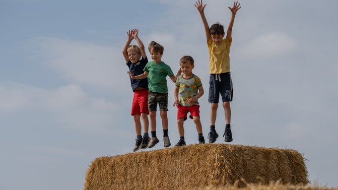 A group of children are standing on the top of a collection of hay bales. It is summer and the sky is bright blue in the background. The children are getting ready to jump off it.