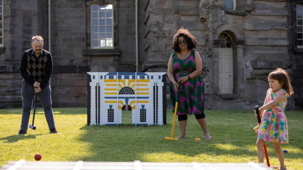 A man, woman and child are holding croquet mallets next to an architectural model with a stone building in the background.