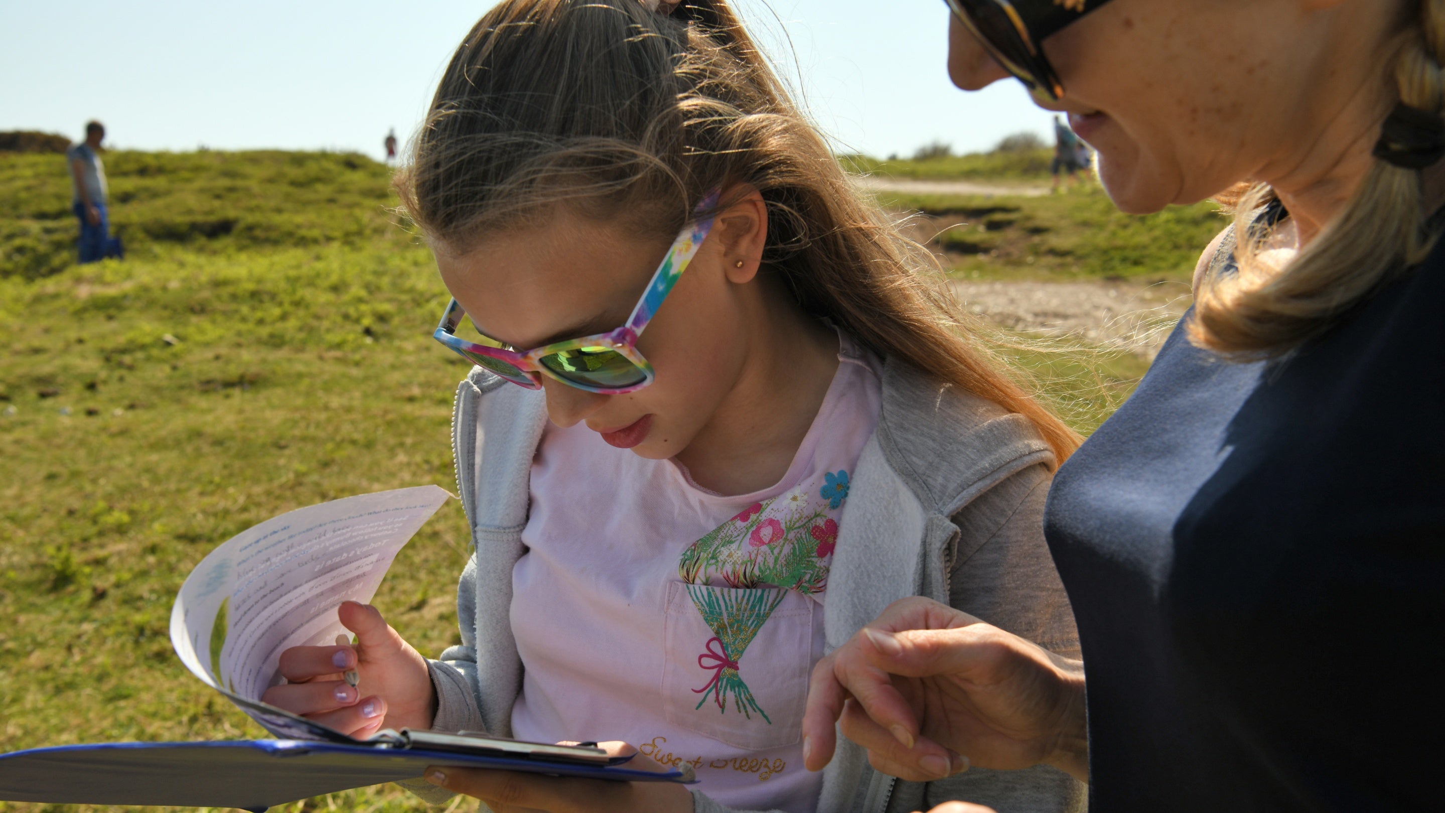 A woman and child stand in sunshine in an open grassy area, both wearing sunglasses and looking at an Easter trail map on a clipboard held by the child