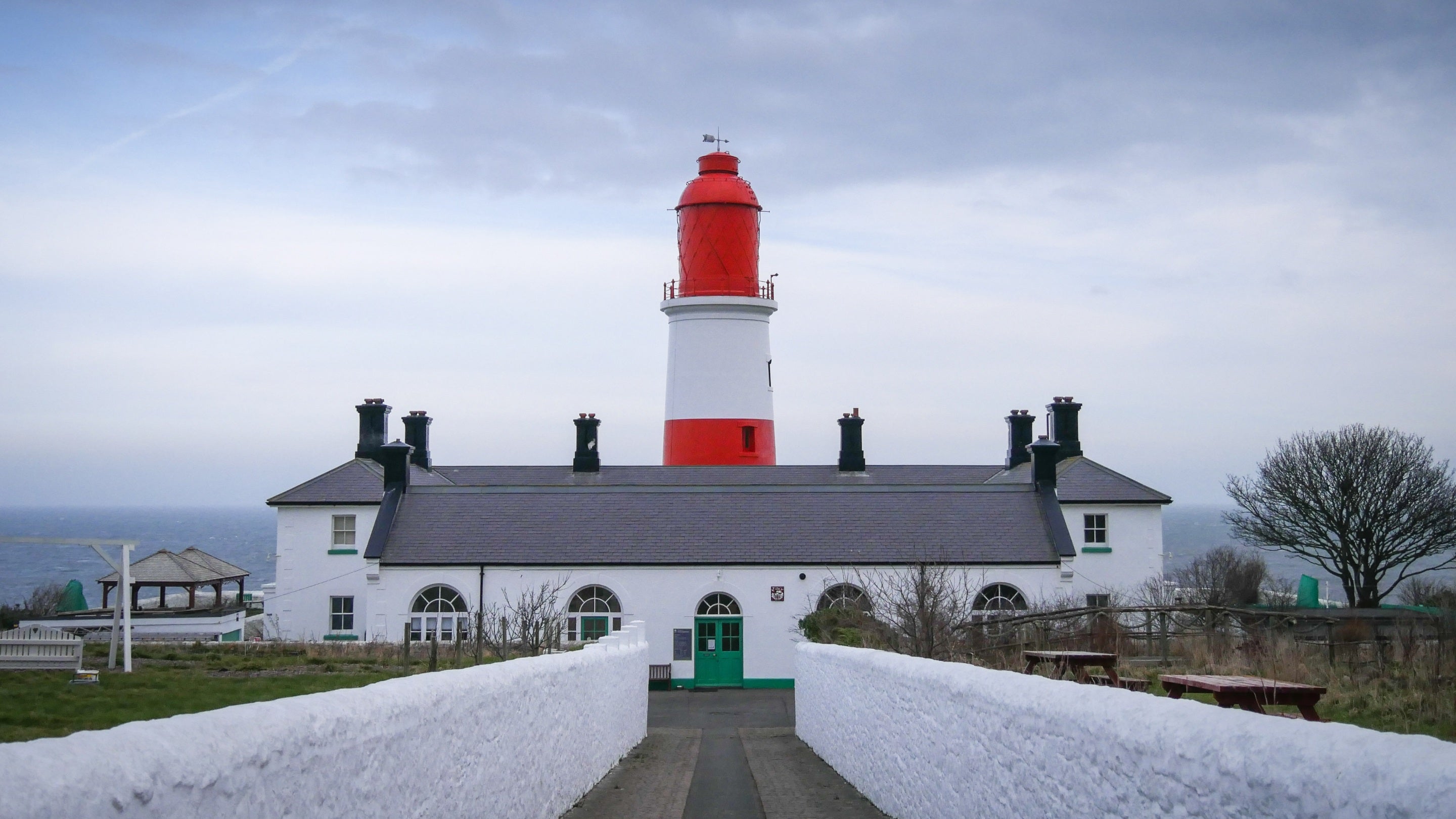 A white walled driveway leads towards the red and white striped tower and white buildings of Souter Lighthouse on a cloudy wintry day.