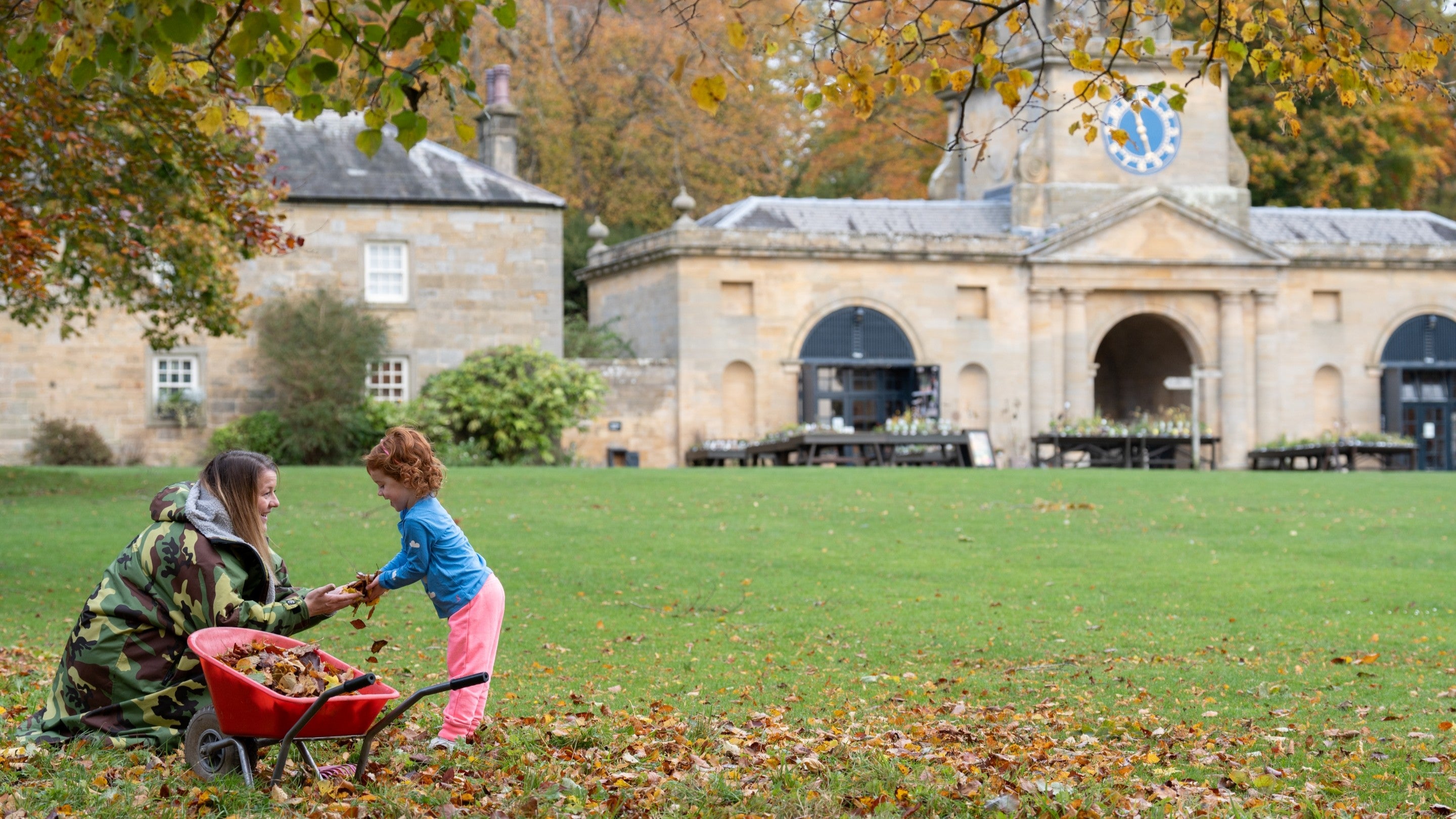 A mum and her daughter putting autumn leaves into a small wheelbarrow. A big clocktower building in the background.