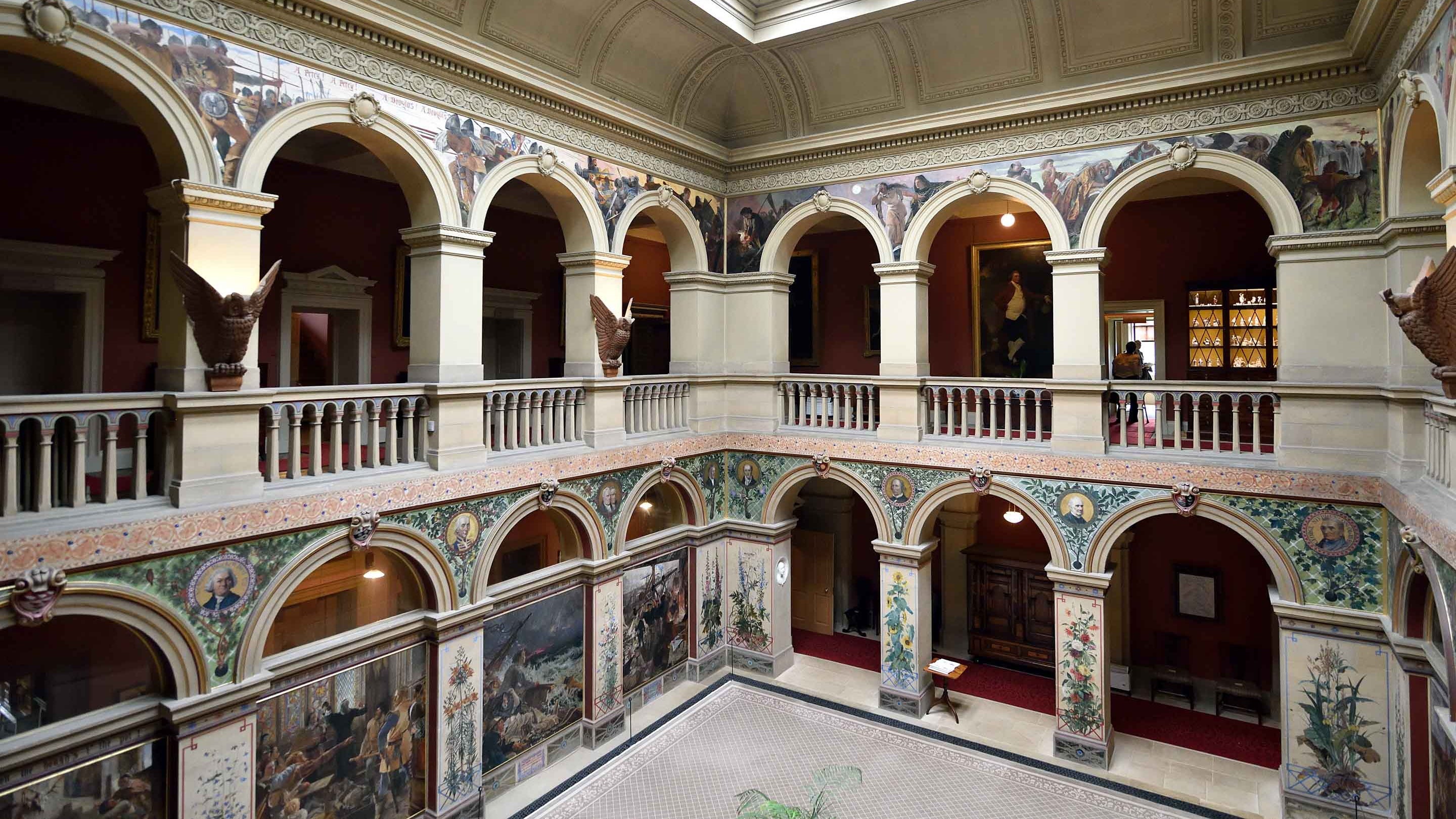 The galleried Central Hall at Wallington, Northumberland, set over two levels with arches between its pillars; the arches are painted with historical figures and scenes.