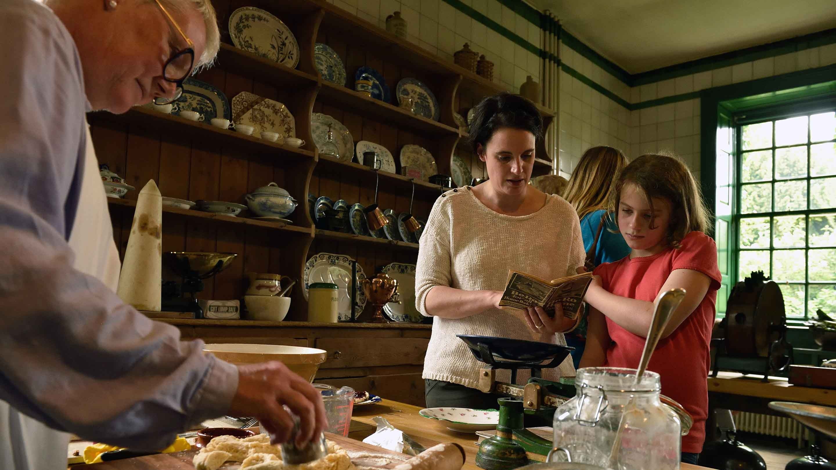 Visitors in the Kitchen at Wallington, Northumberland, look at items on the table, while a volunteer is baking