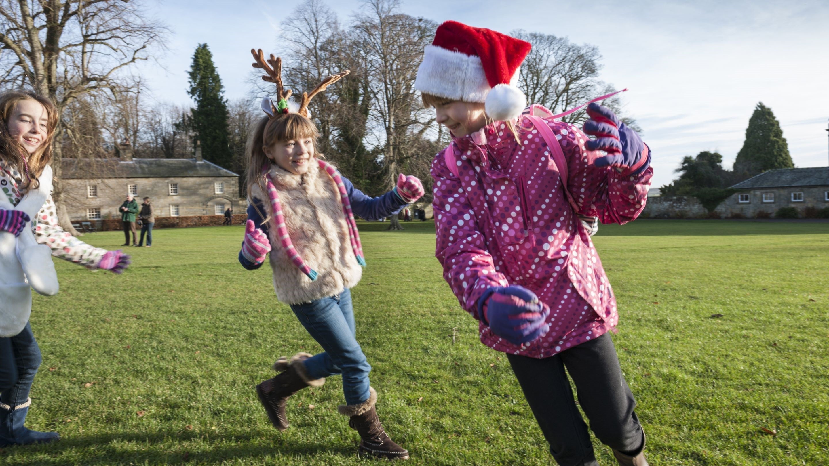 Three girls laughing and running in the grass courtyard at Wallington. One is wearing a Santa hat and another a reindeer headband