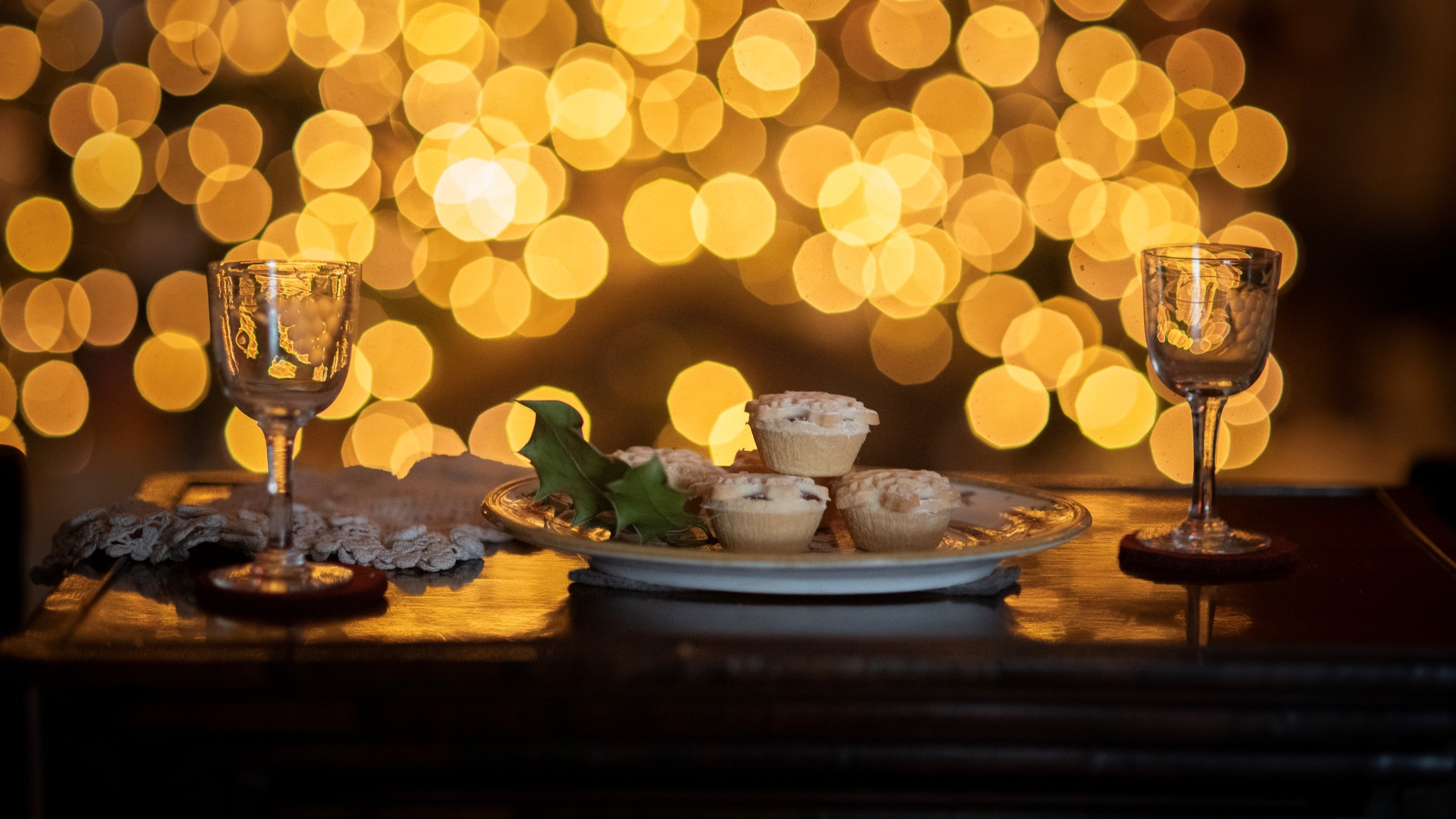 Mince pies on a plate with two small wine glasses on either side and Christmas lights in the background.