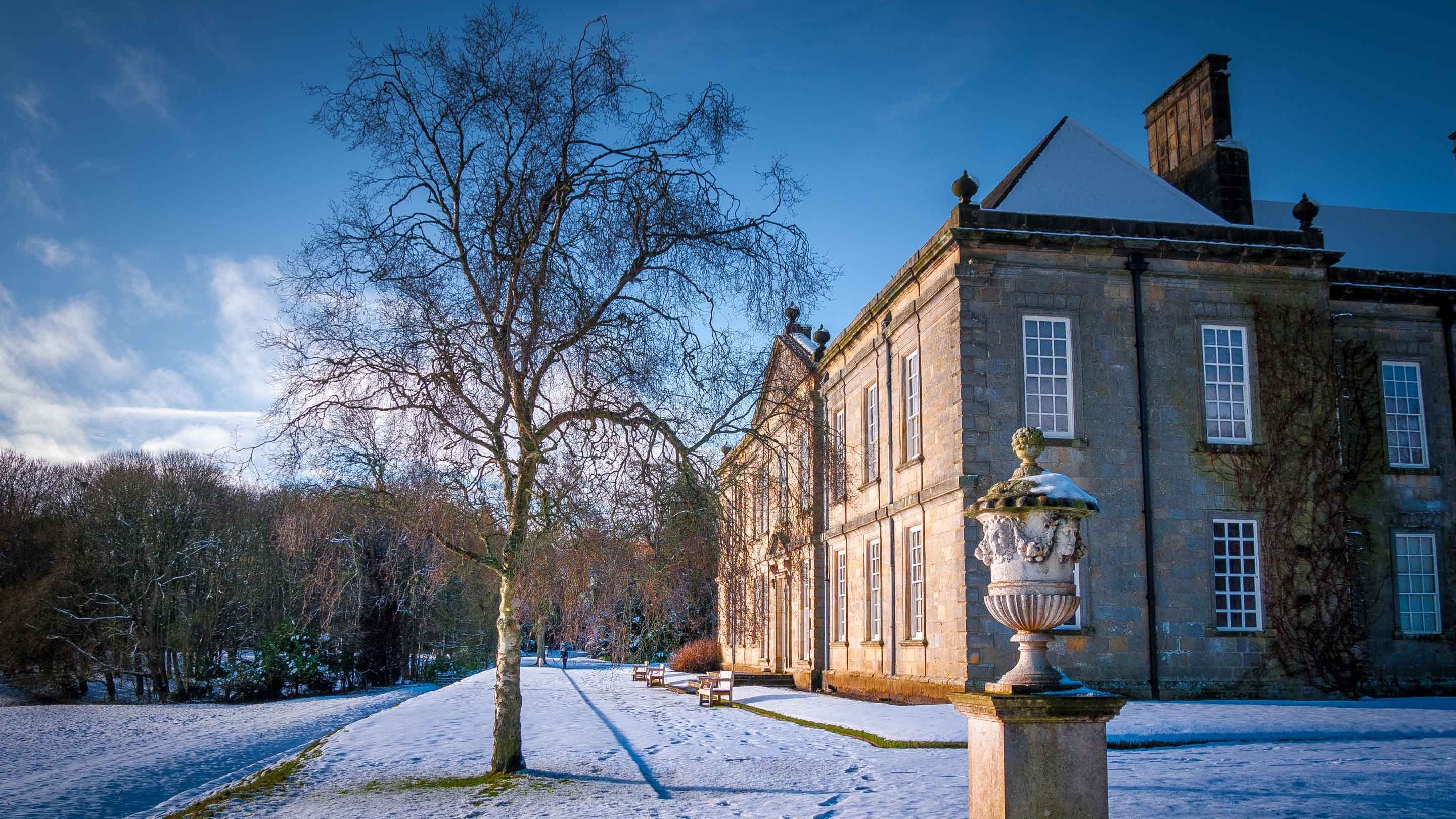 Snow blankets the ground in winter outside the house at Wallington, Northumberland; a large stone urn on a pedestal and a bare tree stand in the foreground.
