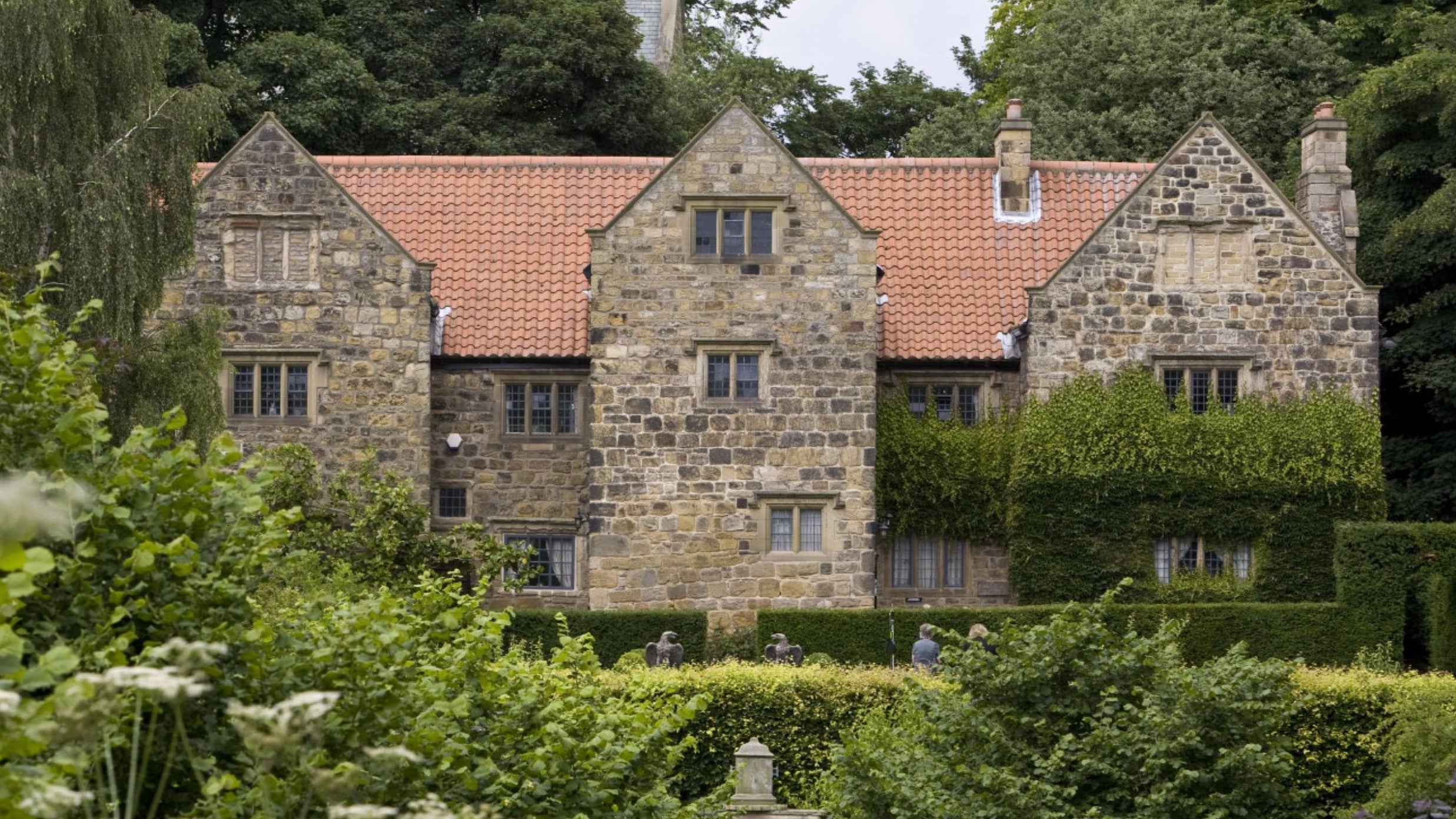 A view of a large stone manor house in the background, with long grasses and hedge in the foreground.