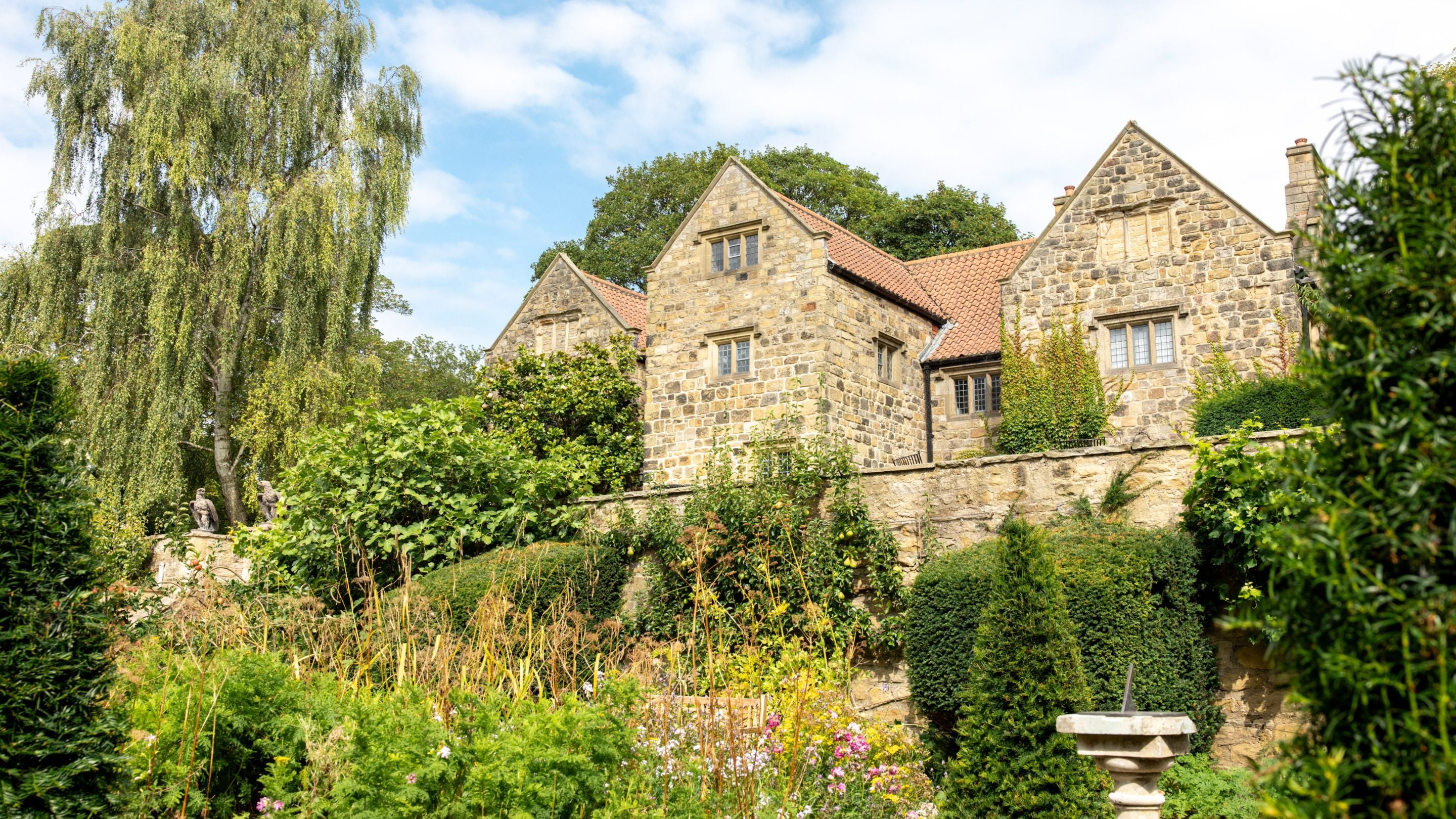 Late summer, blue sky with white clouds. The stone exterior and red tiled roof of Washington Old Hall viewed from gardens below with plants in bloom, topiary shrubs and a stone sundial in the foreground.