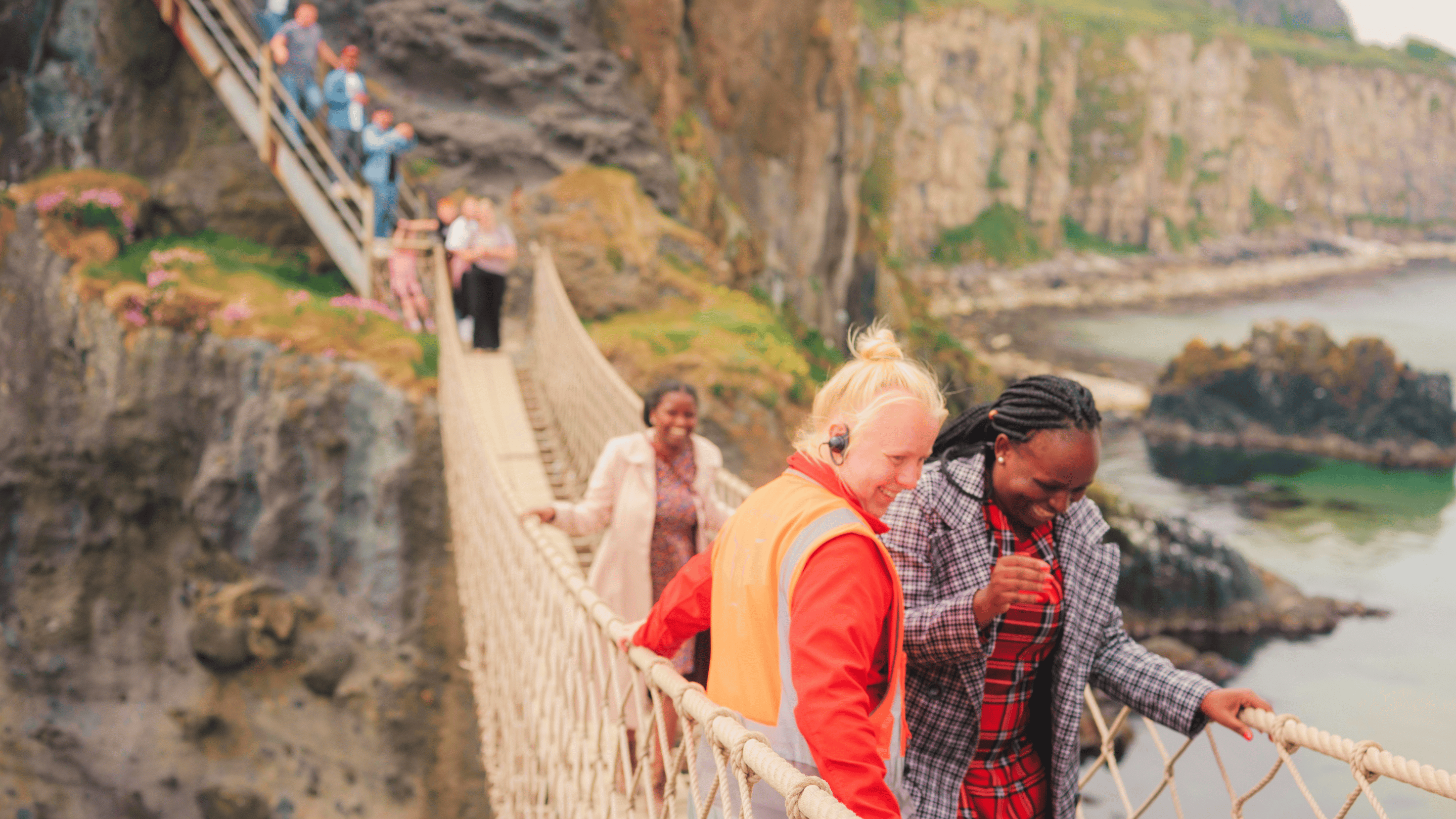 A National Trust team member assisting a visitor, wearing a tartan dress and a long black and white checked jacket, across Carrick-a-Rede Rope Bridge, Co. Antrim. There is a visitor in the background, who's smiling at her friend whilst she walks across onto the island from the bridge