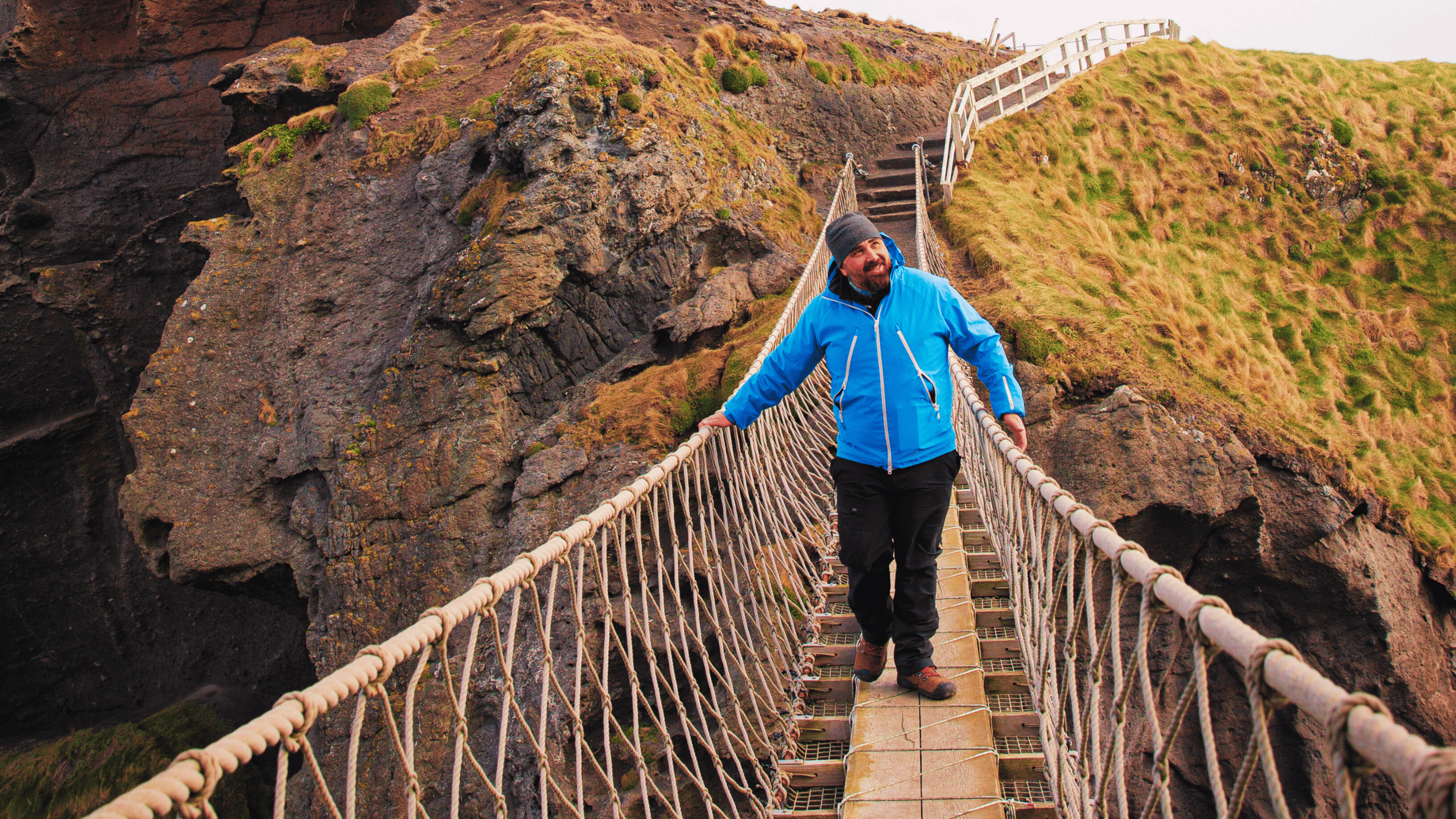 A visitor wearing a blue waterproof jacket, black outdoor trousers and a grey hat, walking across Carrick-a-Rede Rope Bridge and admiring the views of the surrounding cliffs