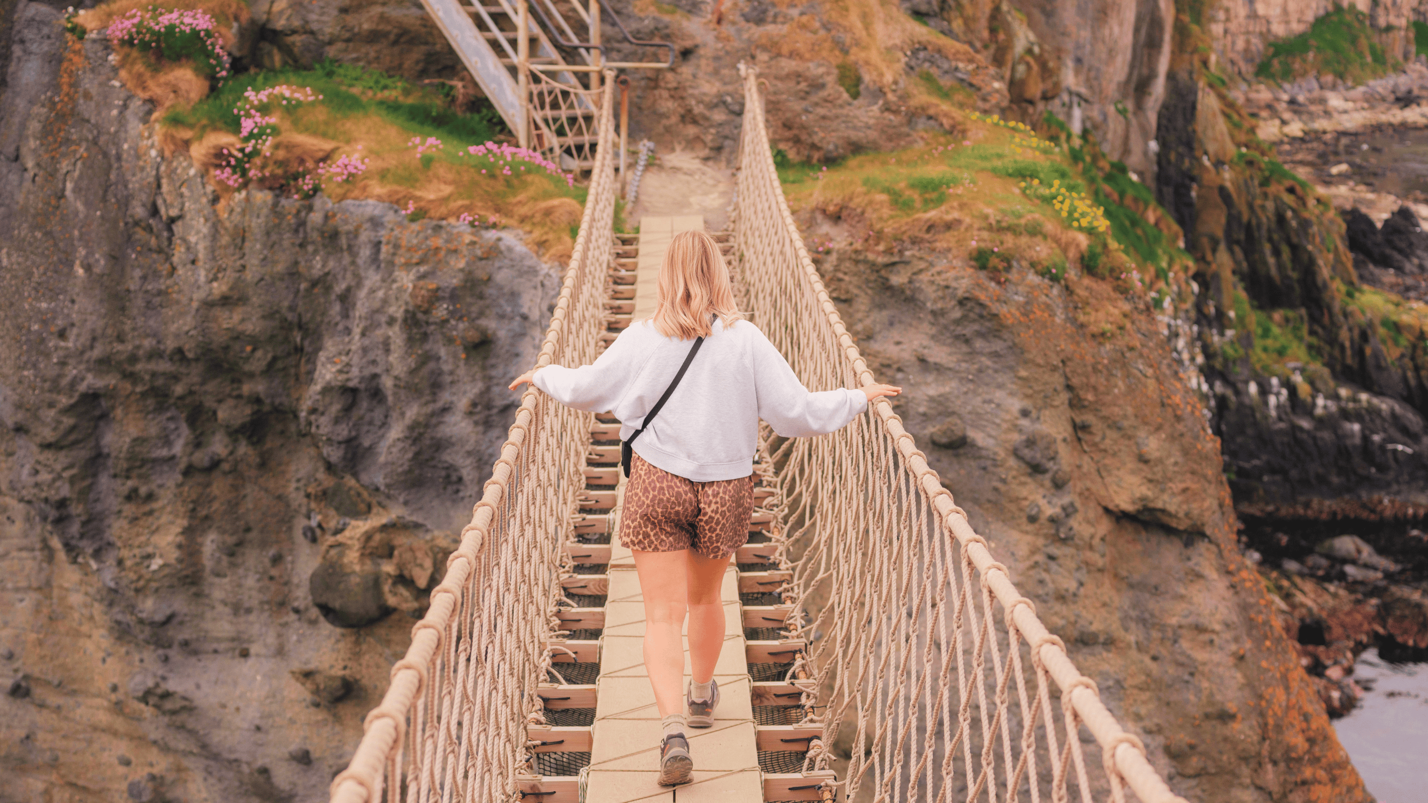A visitor wearing a grey jumper and leopard print shorts, walking across Carrick-a-Rede Rope Bridge, captured from the island
