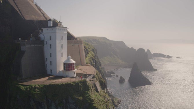 White lighthouse on Rathlin Island, with a red and white lantern room perched on a steep, green cliff overlooking the ocean, with rocky formations below and an overcast sky above