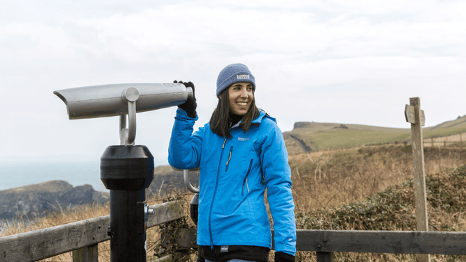 Visitor in a blue rain coat and beanie hat, about to look out through a telescope on the mainland at Carrick-a-Rede, out towards Rathlin and the Scottish islands