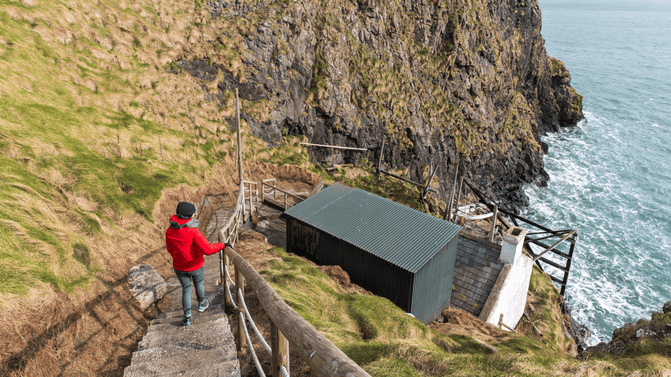 Visitor with a red rain coat, blue jeans and blue/black hiking boots, walking down the stone steps towards the Fisherman's Cottage on Carrick-a-Rede island