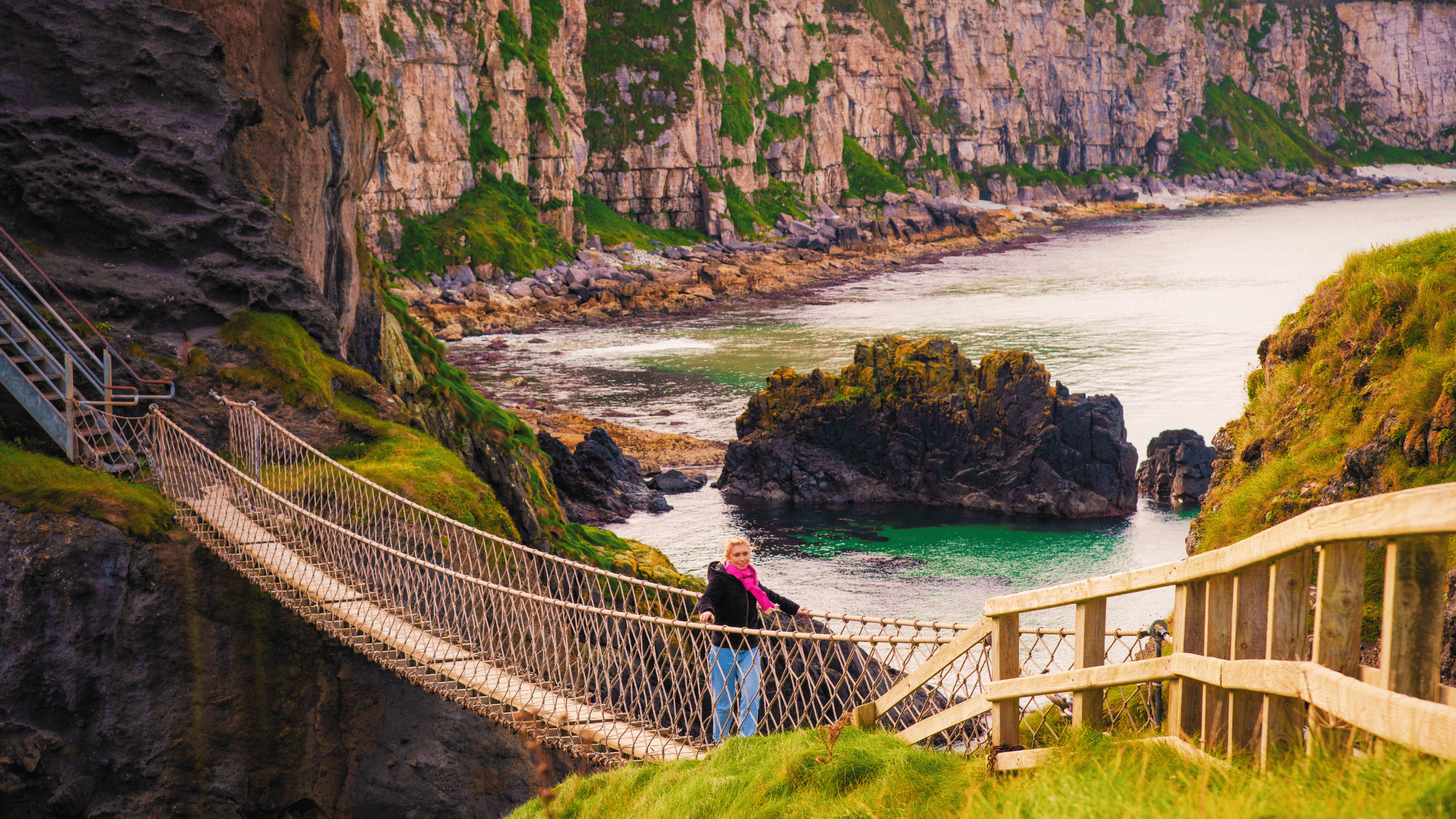 Visitor crossing Carrick-a-Rede Rope Bridge on an autumn day, with rugged coastal cliffs, rocky outcrops and calm turquoise waters below