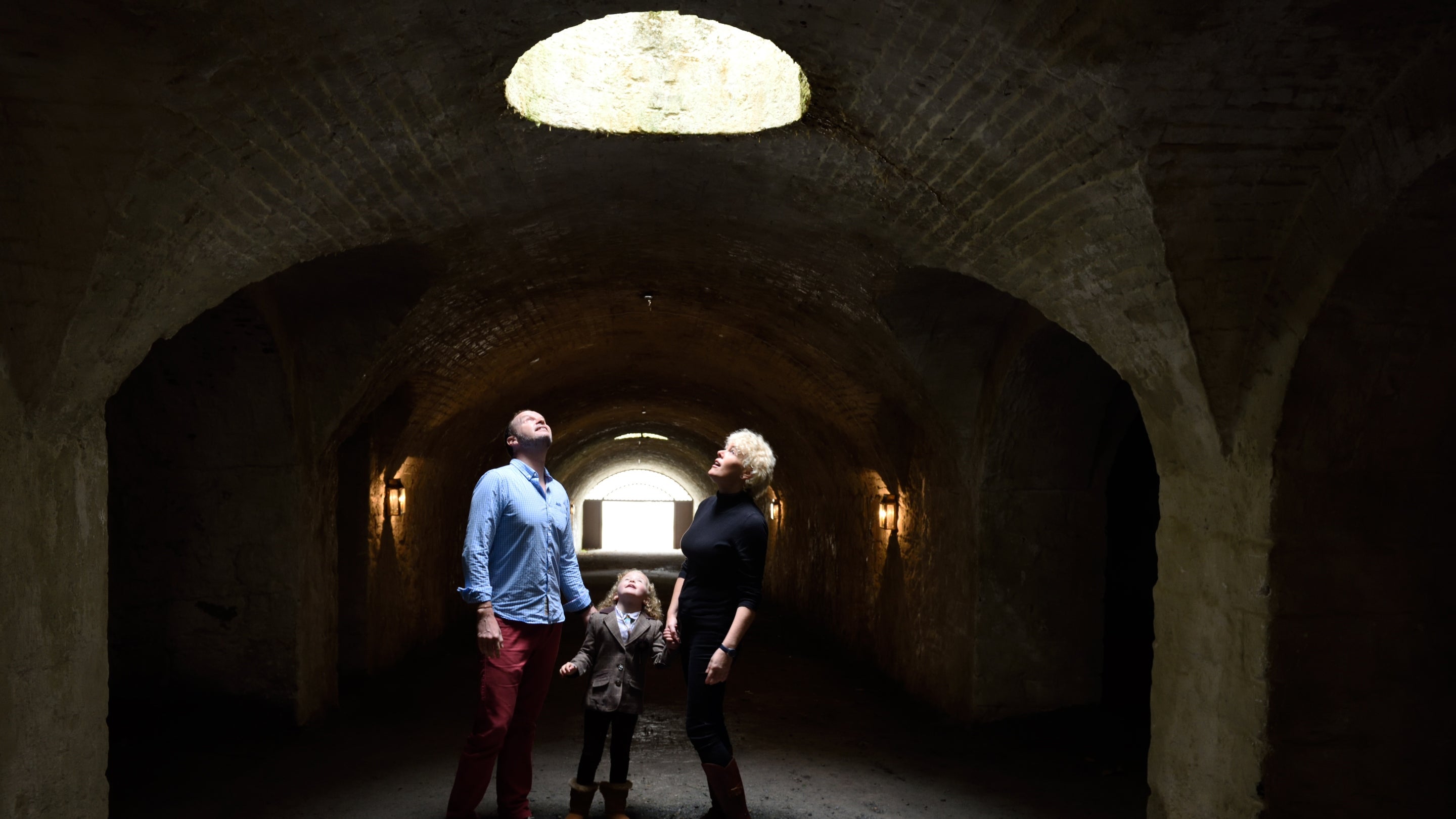 A man, woman and child in a dark tunnel looking up at a light coming through a hole in the ceiling