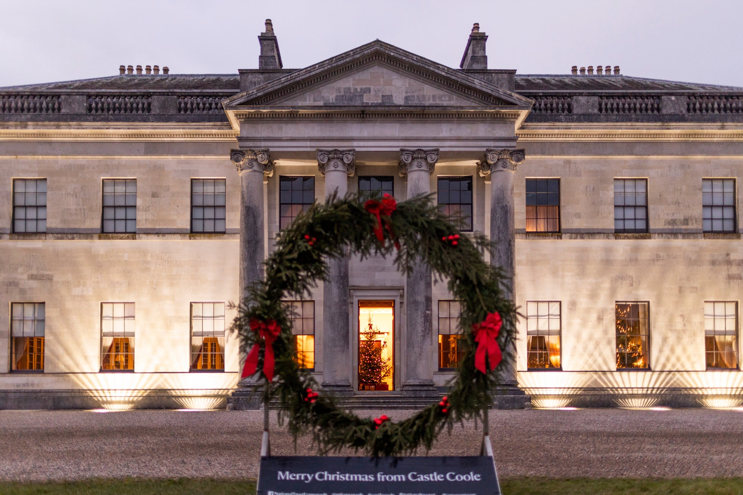 Castle Coole mansion lit up at nighttime with a wreath in front saying Merry Christmas from Castle Coole