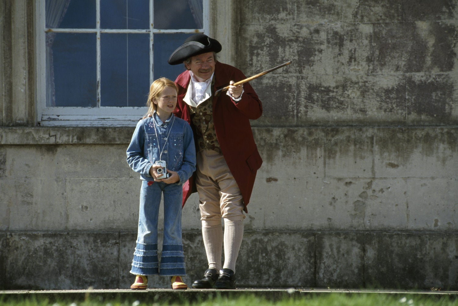 A small girl with a man in c18th costume, he is pointing out something to her with a cane, in front of the house at Castle Ward