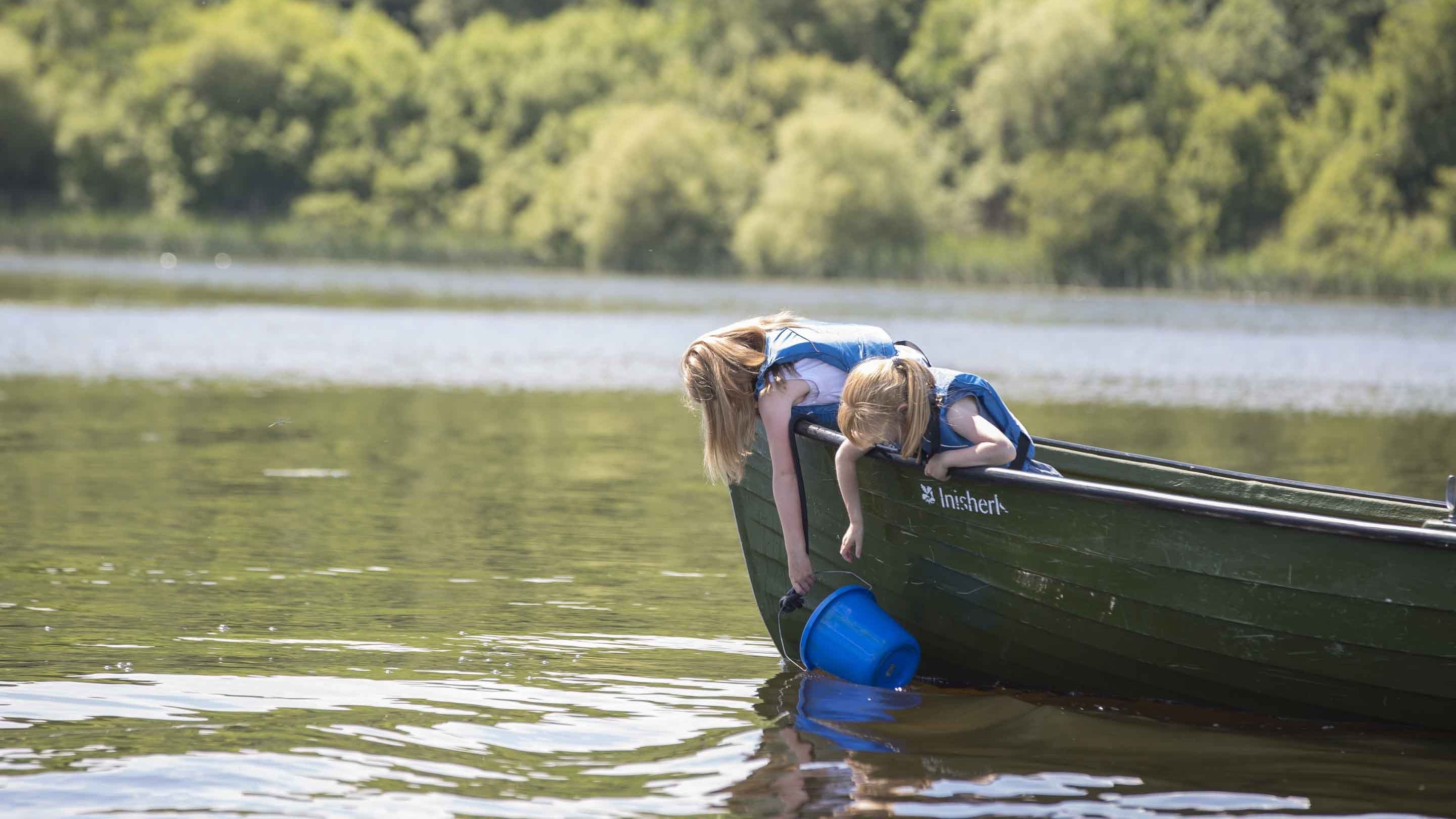 Two children leaning over the side of a boat with a bucket in the water of Lough Erne at Crom, with trees in the background and a stretch of the river visible