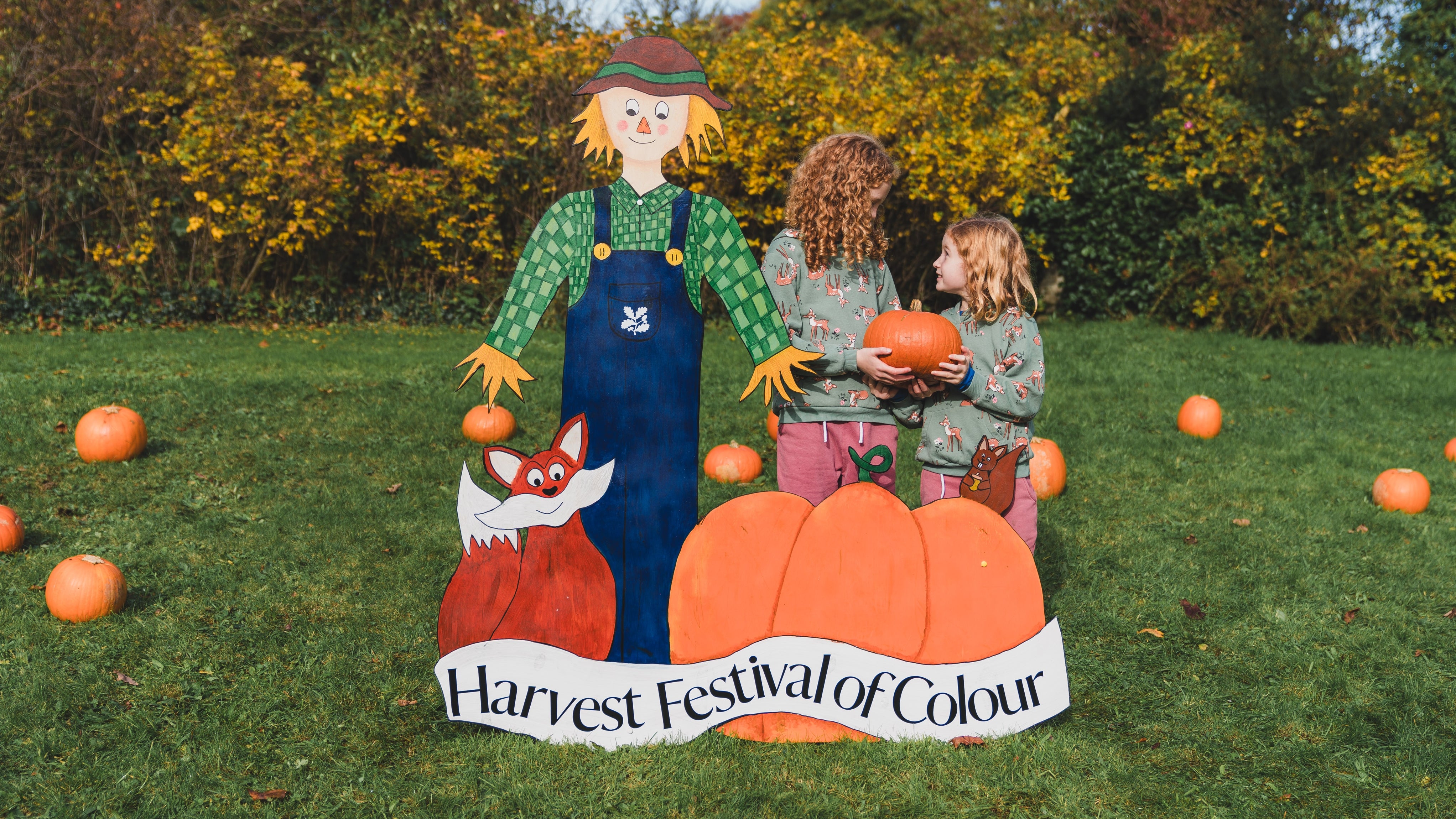 Two sisters holding pumpkins beside a sign saying Harvest Festival of Colour