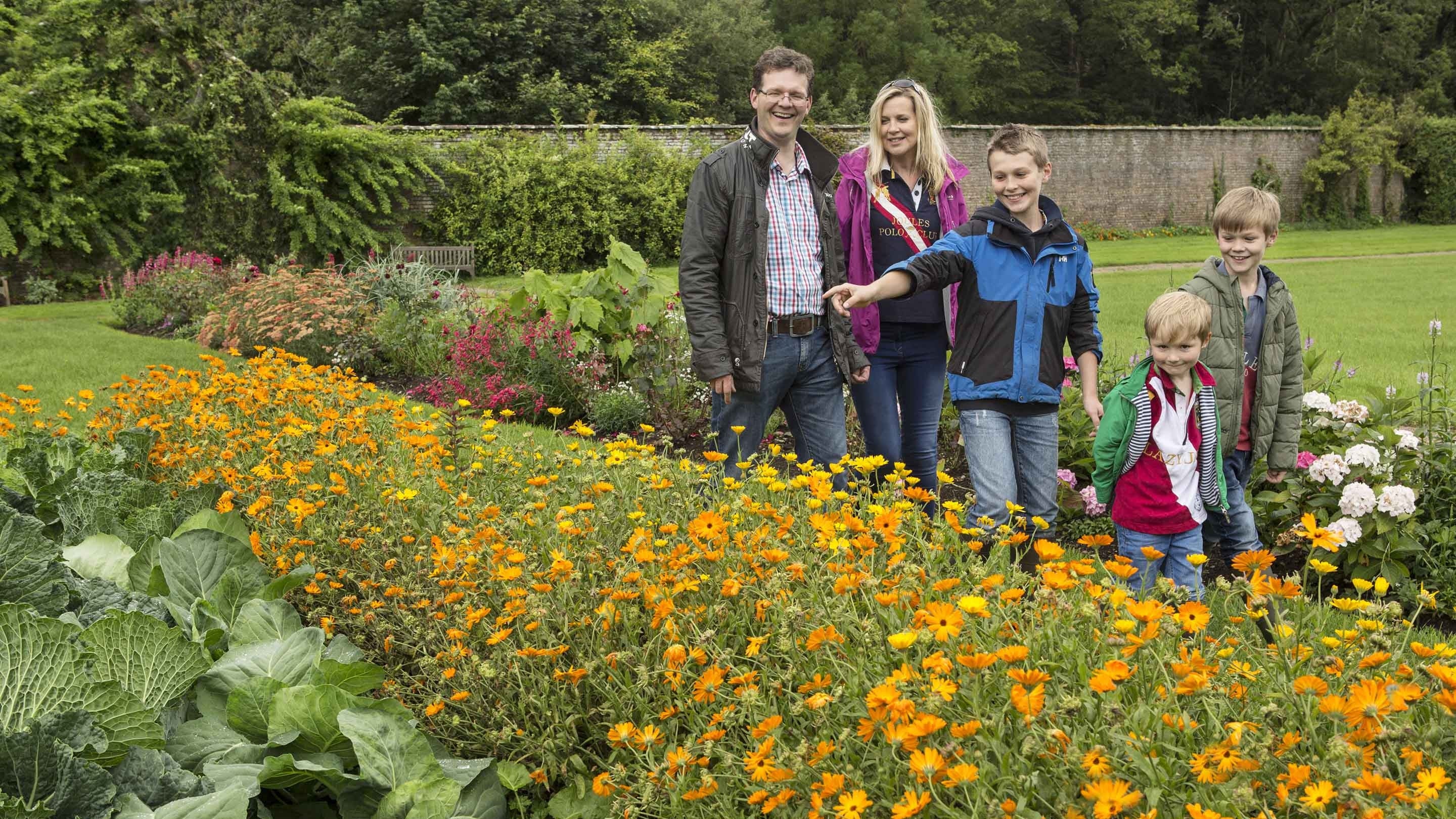 Two adults and three children are walking between flowerbeds at Florence Court, County Fermanagh. They are all smiling and one child is pointing at a flowerbed blooming with orange flowers.