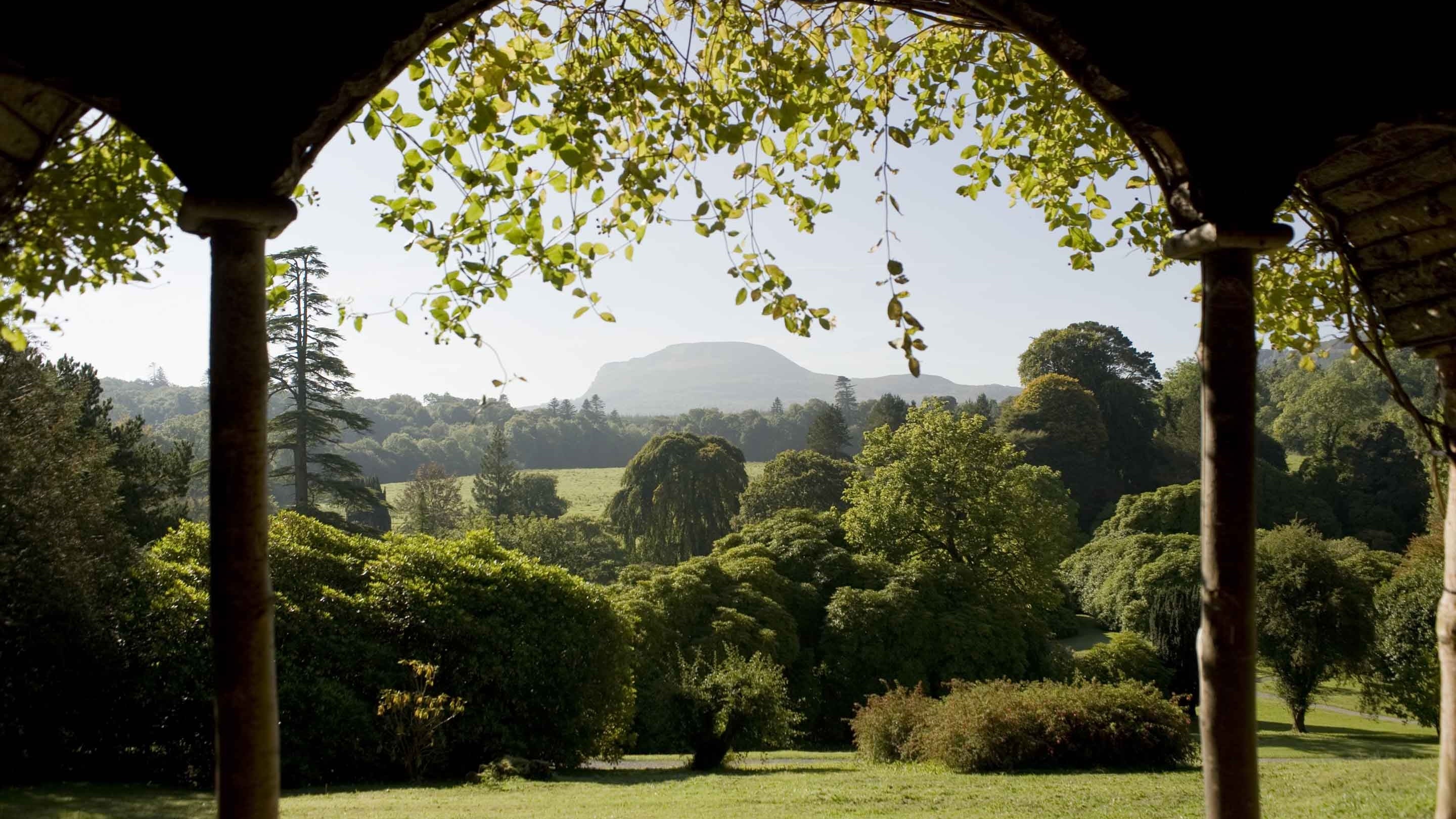 A view through the pillars of the Summer House at Florence Court, County Fermanagh, looking out over the lawn and mature trees in the garden and towards the surrounding countryside.