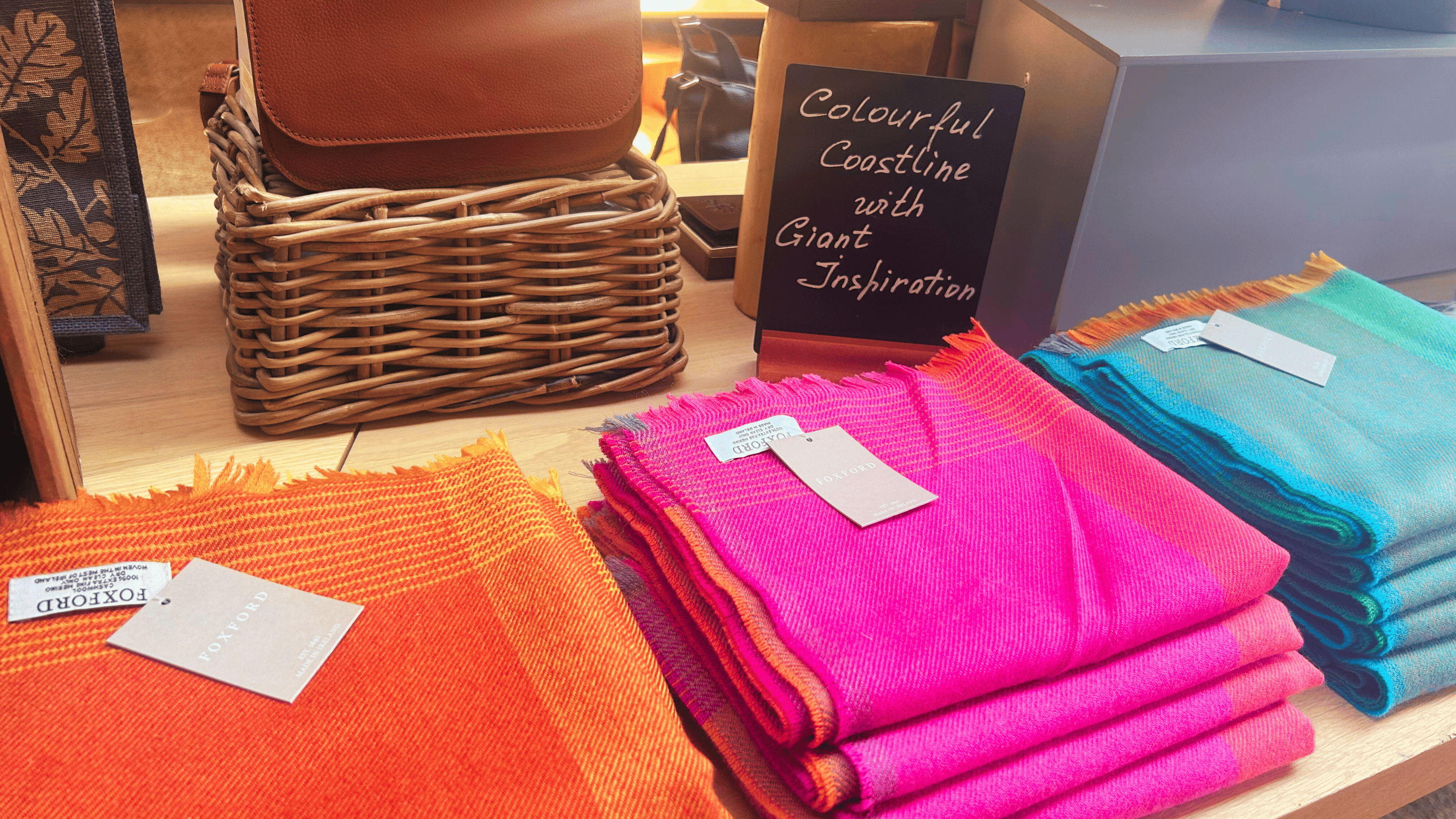 3 multi-coloured Foxford scarfs lined in display in Giant's Causeway Visitor Centre Retail area. Wicker basket at the back, with a brown satchel on top and a black chalkboard sign that states, 'Colourful coastline with giant inspiration'.