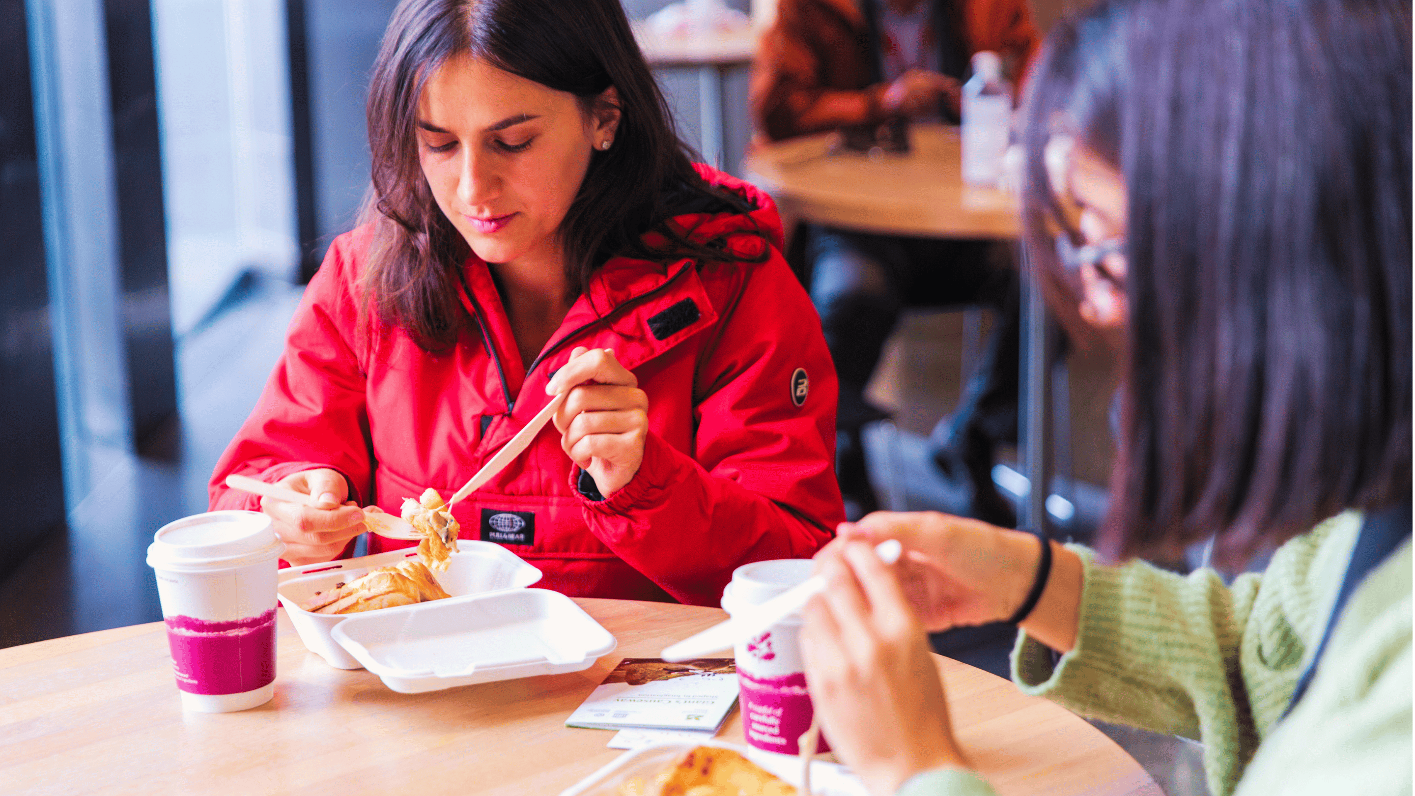 2 visitors enjoying a meal at a table in the Giant's Causeway Visitor Centre Café, with takeaway coffee cups and food containers in a bright indoor seating area and other café guests sitting in the background