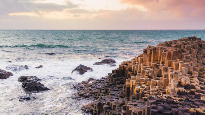 View of the Giant's Causeway with waves crashing against hexagonal basalt columns under a partly cloudy sky at sunrise or sunset