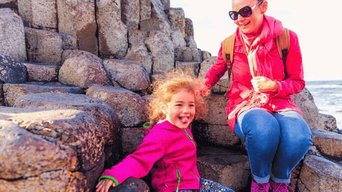 An adult and child sit on the Giant's Causeway basalt columns, with the child seated on the famous Wishing Chair and the Atlantic Ocean visible in the background