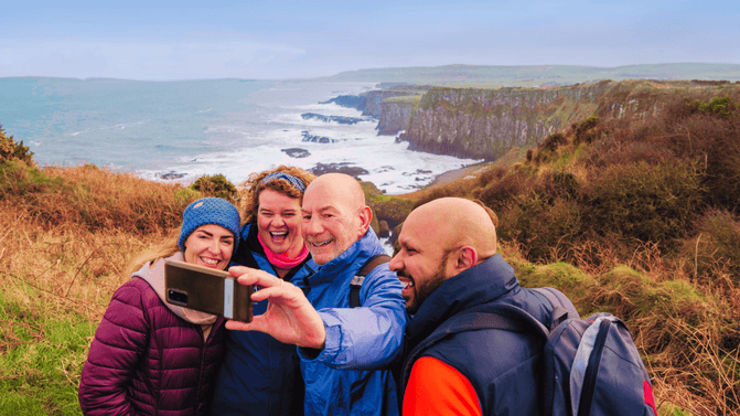 Three people taking a selfie on the clifftops overlooking the rugged cliffs and waves of the Causeway Coast in Northern Ireland