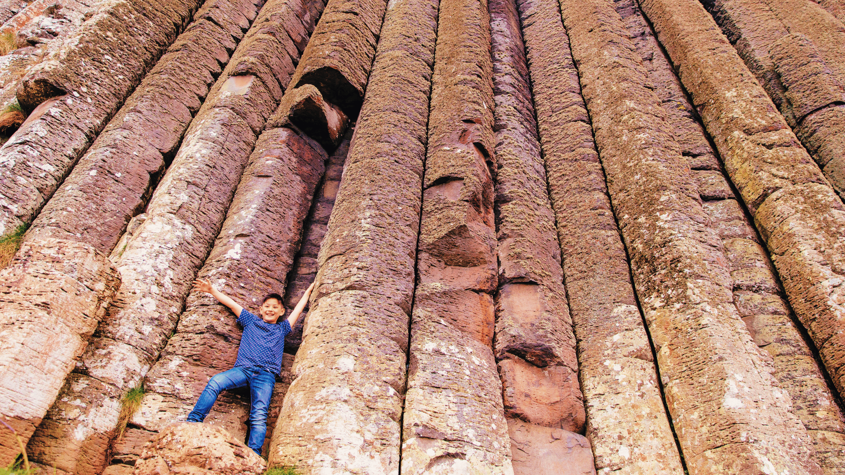 A young visitor with their hands in the air, standing on the base of a long trunk-like rock (otherwise known as the Amphitheatre) at the Giant's Causeway, Co. Antrim