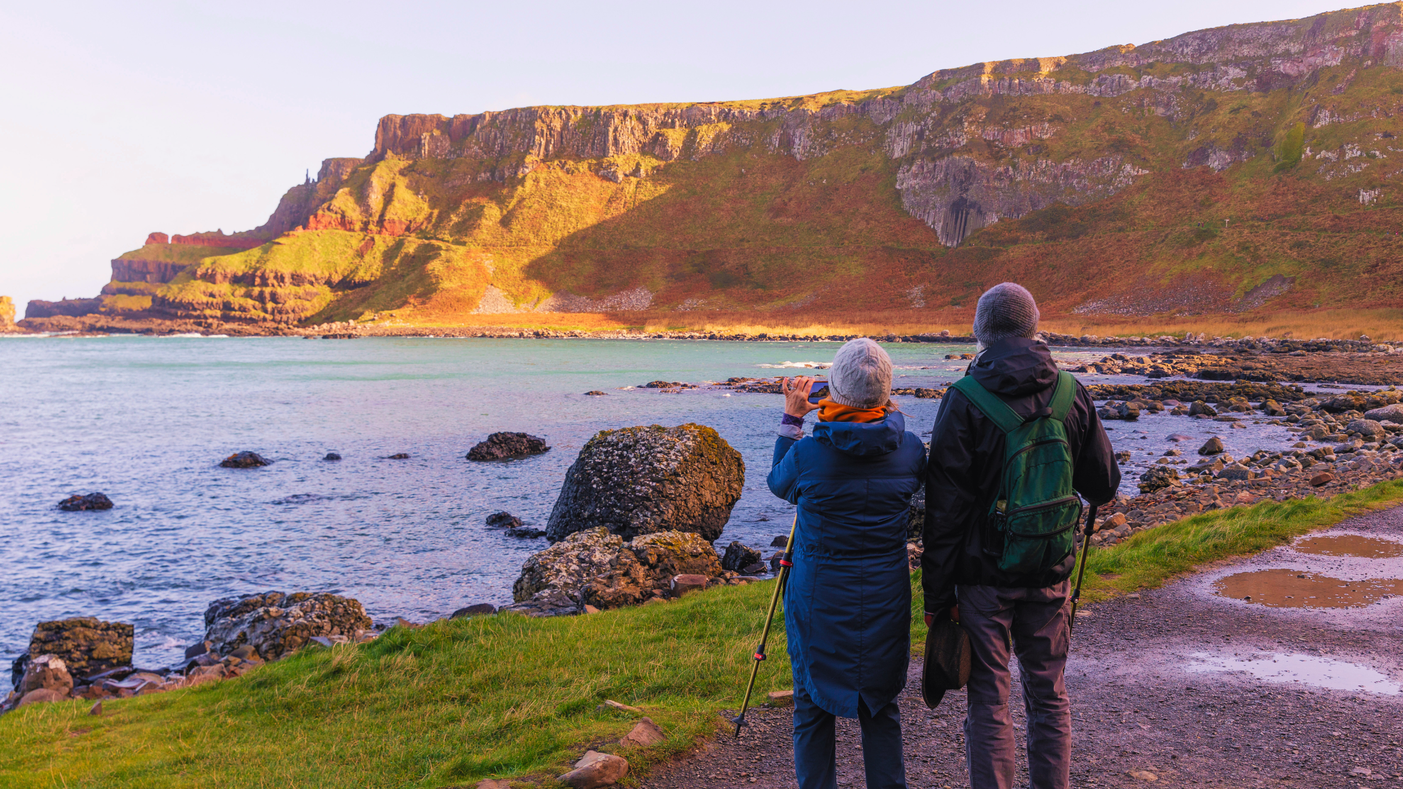 Two visitors admiring the Causeway Cliffs from Port Noffer, with one taking a picture on their phone. Both visitors are wrapped up warm in long coats, hats and scarfs and are both holding hiking sticks.