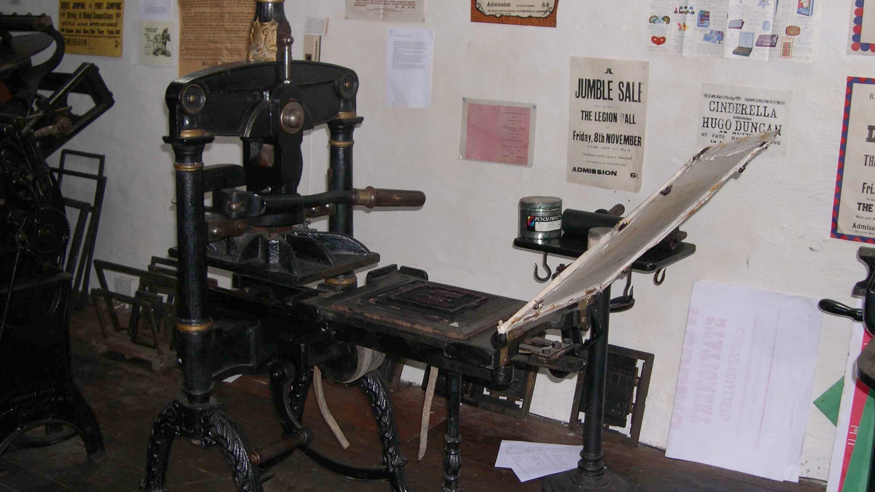 The small Albion Press at Gray's Printing Press, set in front of a wall covered in printed posters and leaflets.