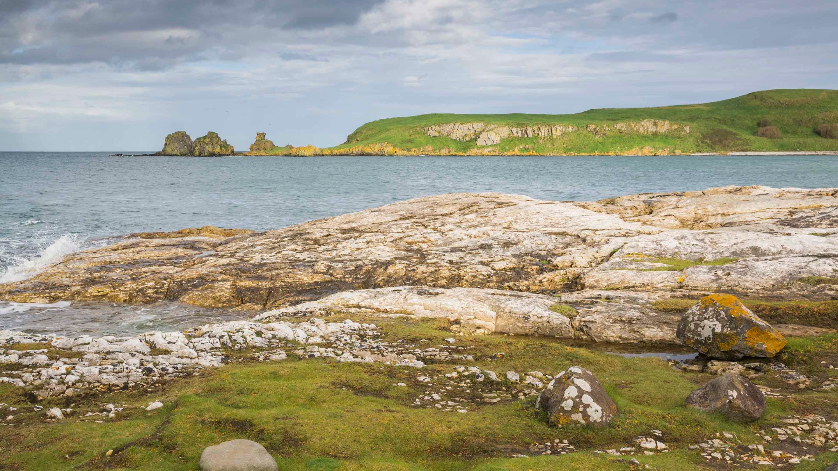 Lichen-covered rocks pepper the grass leading to a large rocky section of the Portmuck coastline on the Islandmagee peninsula, Country Antrim, Northern Ireland. A grassy headland is visible in the distance.