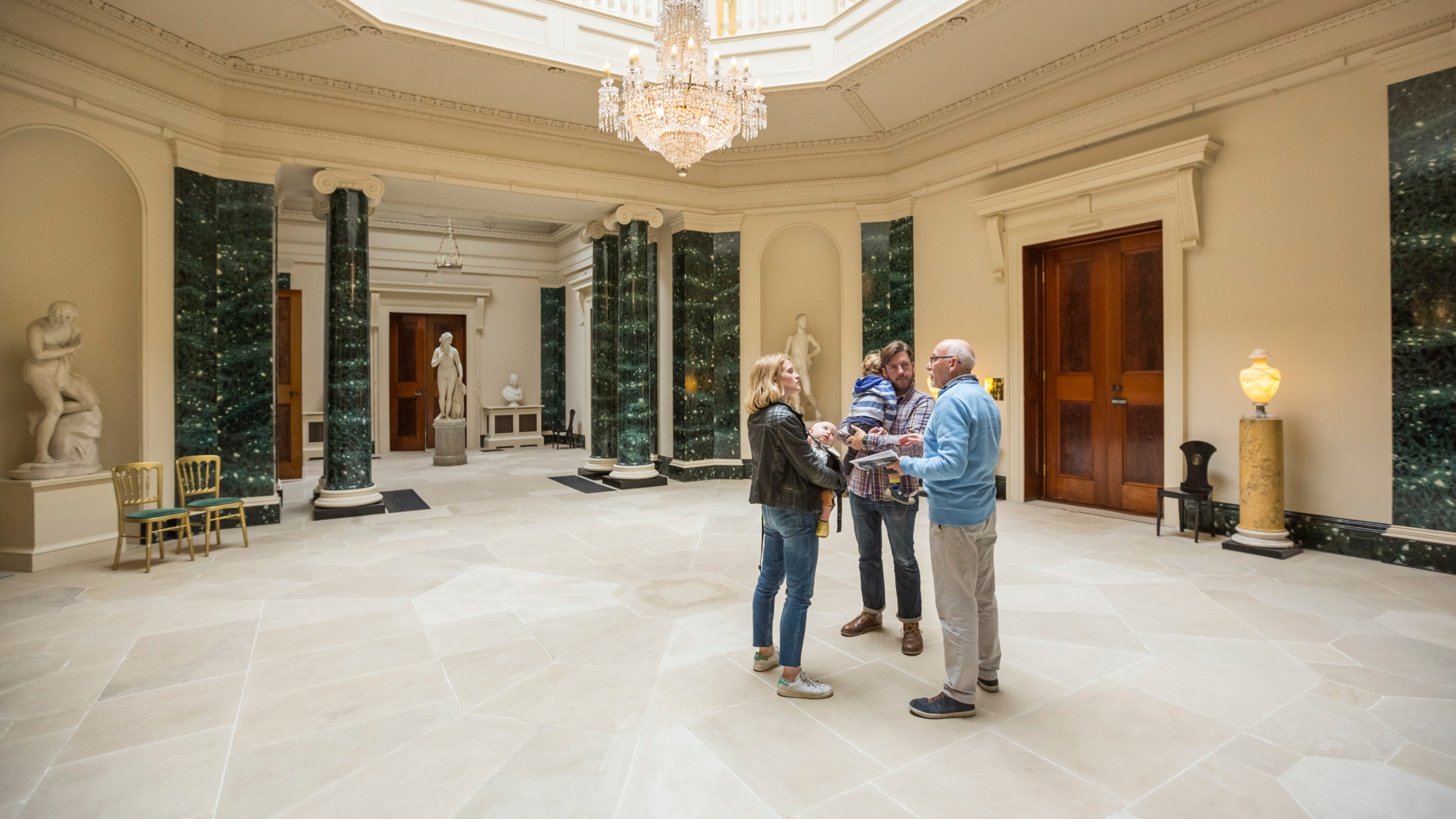 A family with small children talking to a volunteer room guide in Central Hall at Mount Stewart, with its stone floor, statues and chandelier visible
