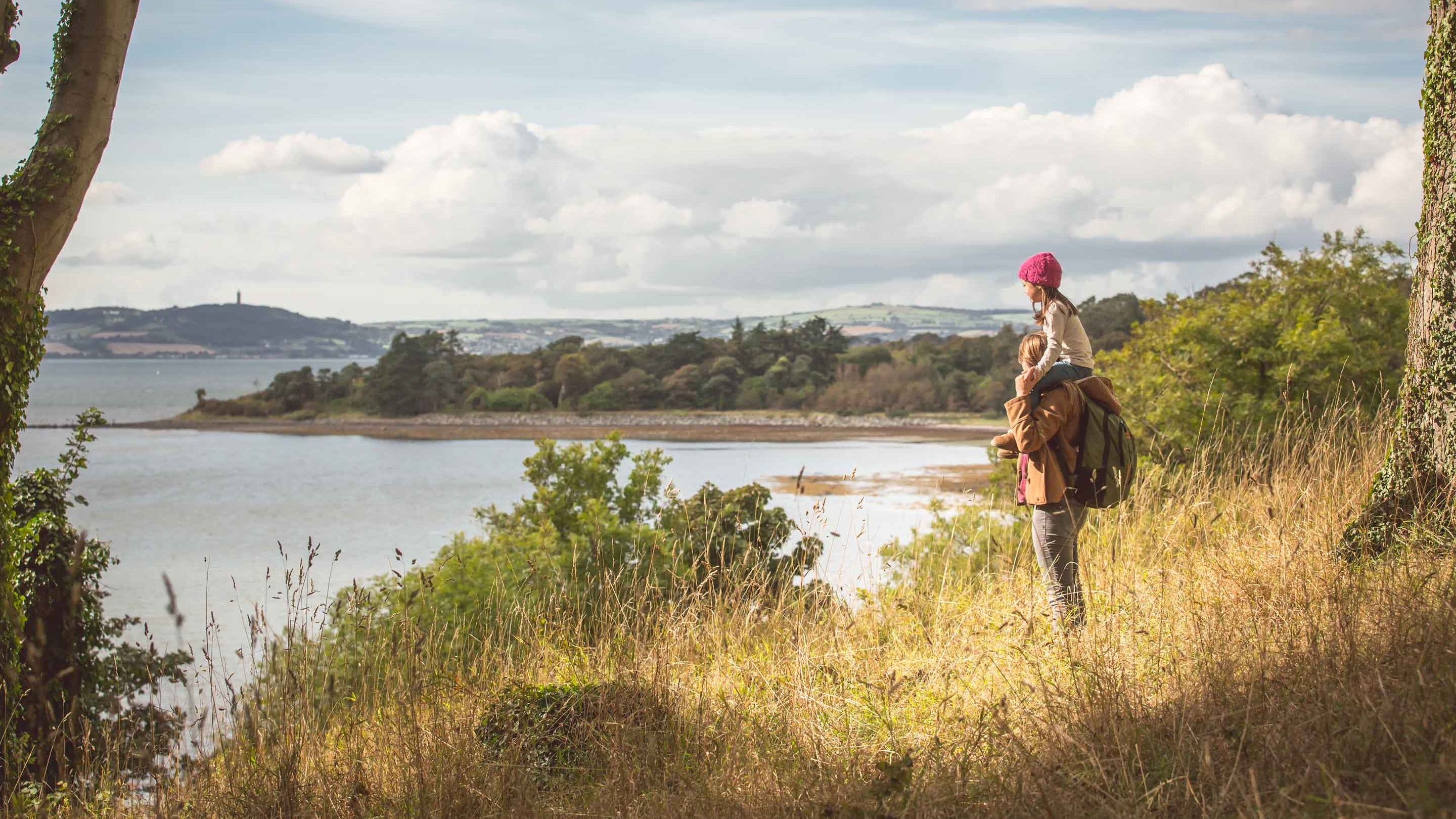 A child on their parent's shoulders admiring a view across the water of Stangford Lough from Mount Stewart