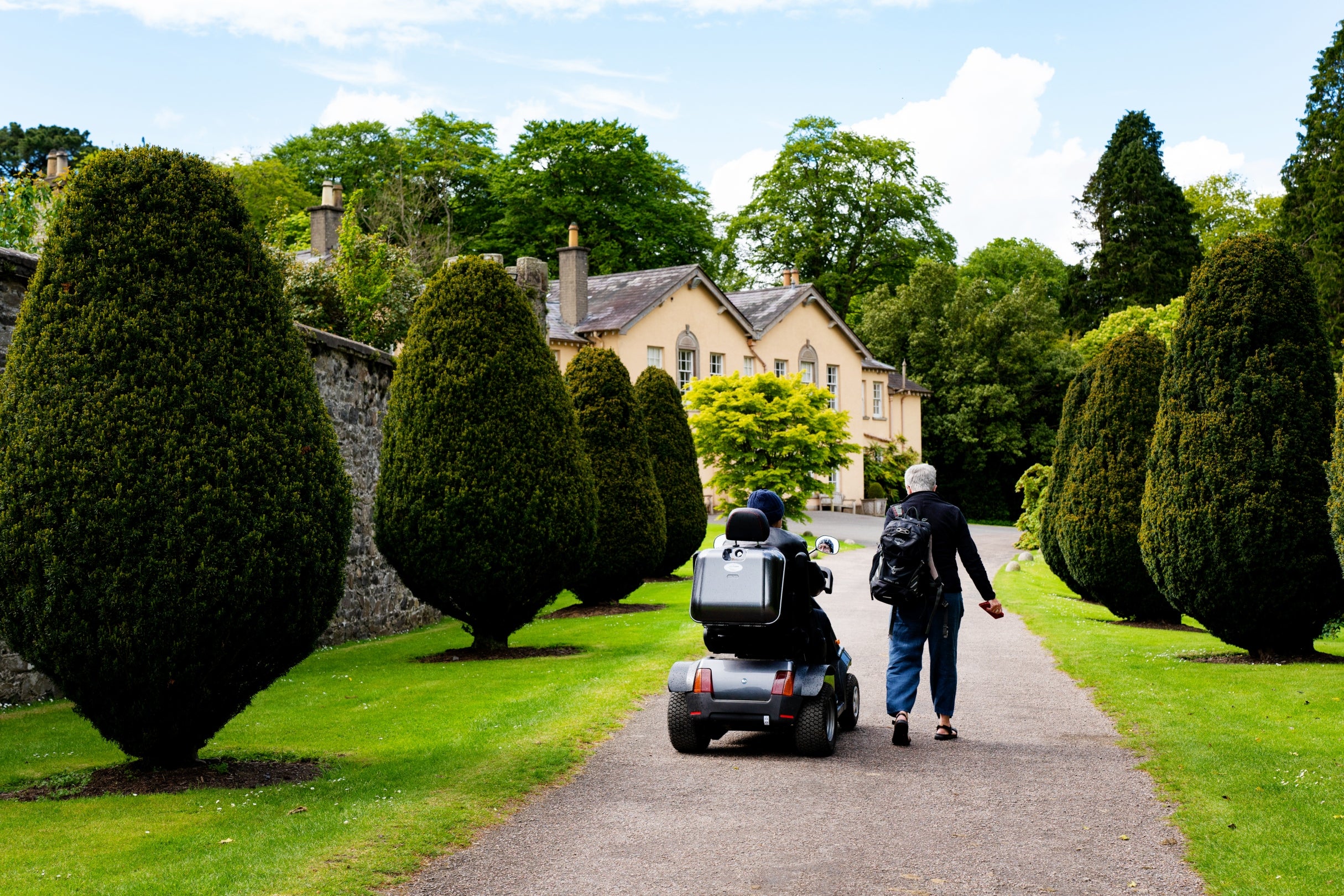 A couple visiting Rowallane Garden, one is in a mobility scooter, travel up the Yew Tree avenue