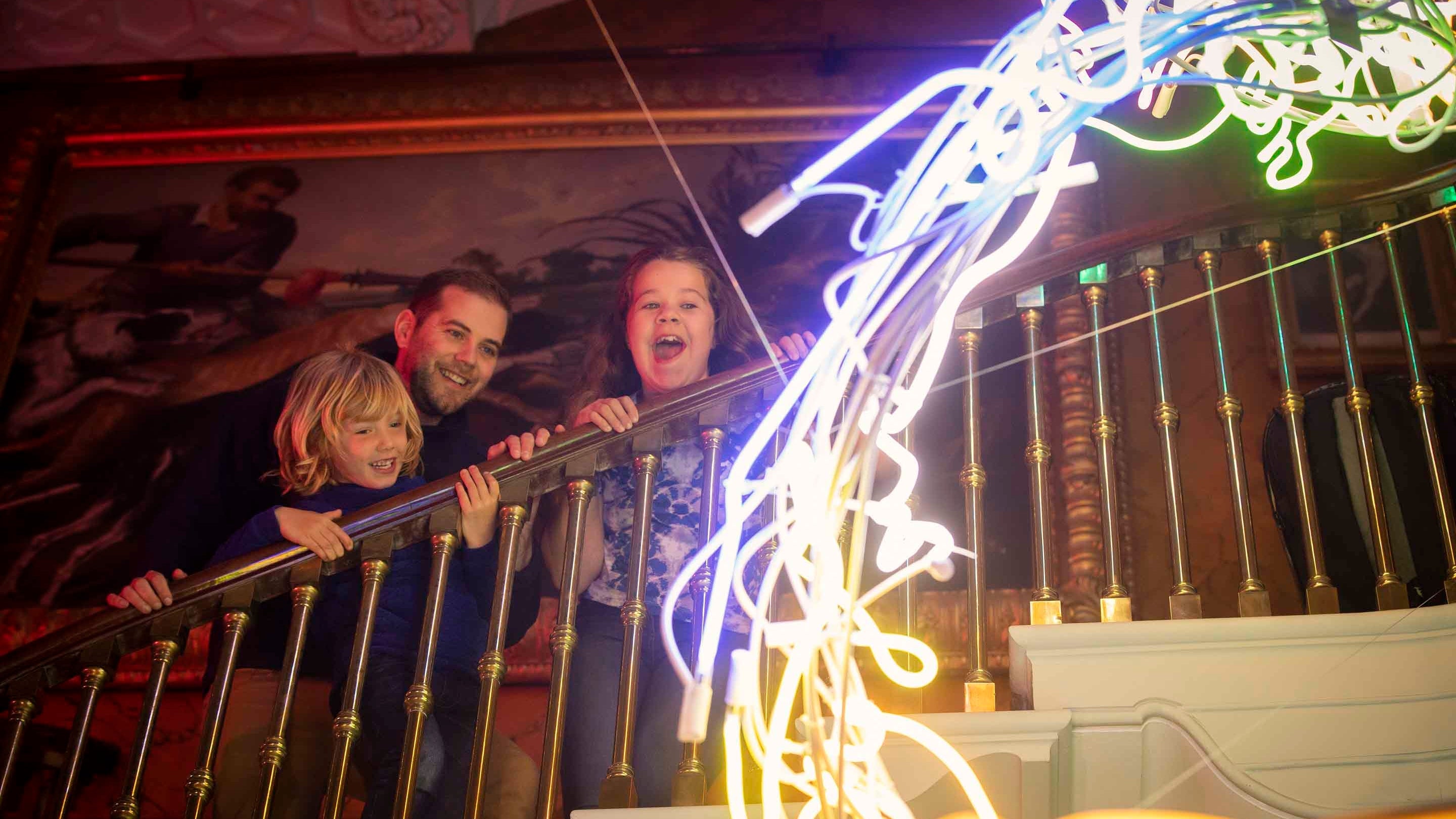 Family admiring the Artifical Sunshine neon sculpture installation in the West Hall at The Argory, with the family standing on a staircase with the neon sculpture in the foreground