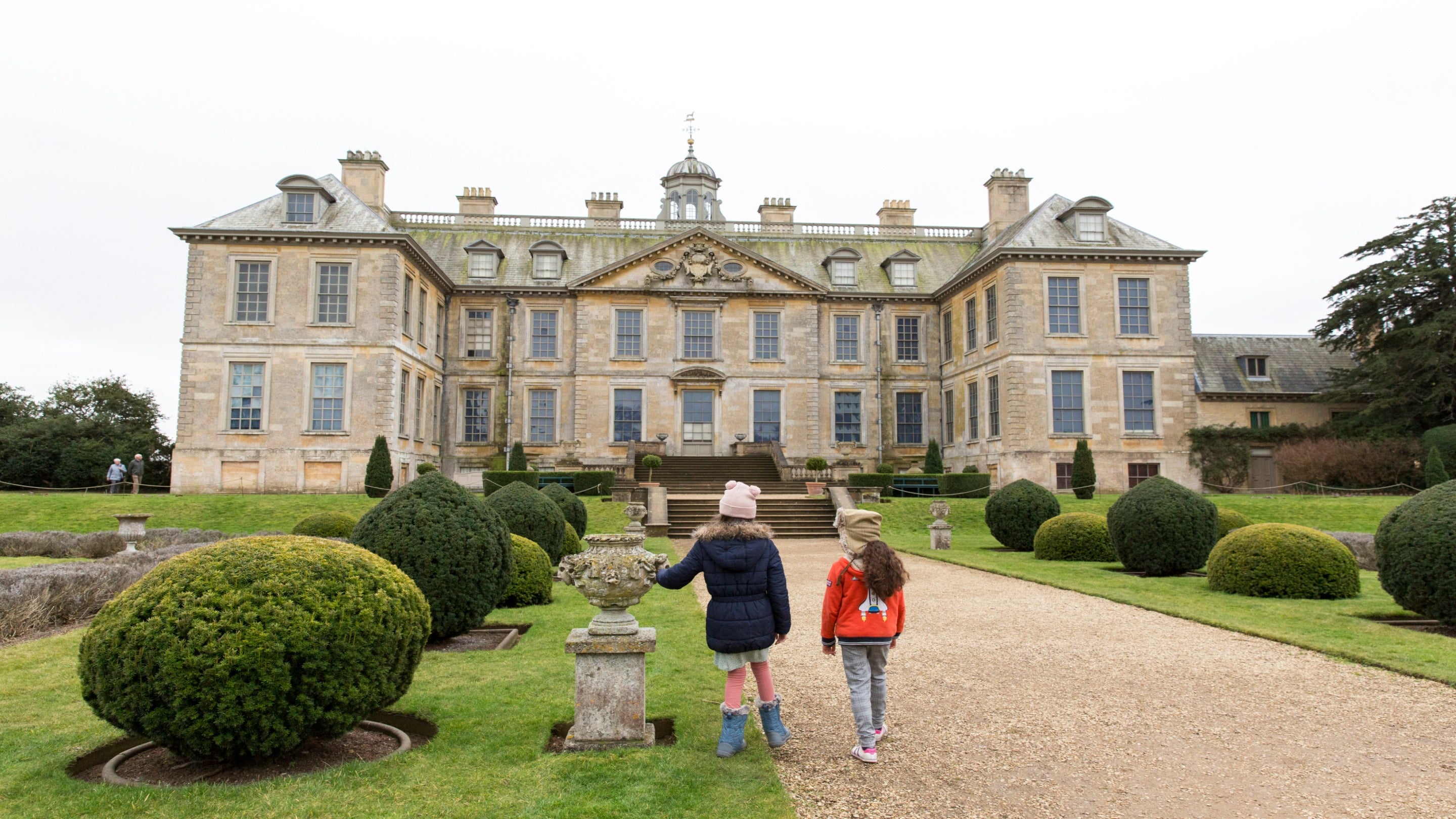 Two young girls in coats and hats walking up gravel park to Belton House