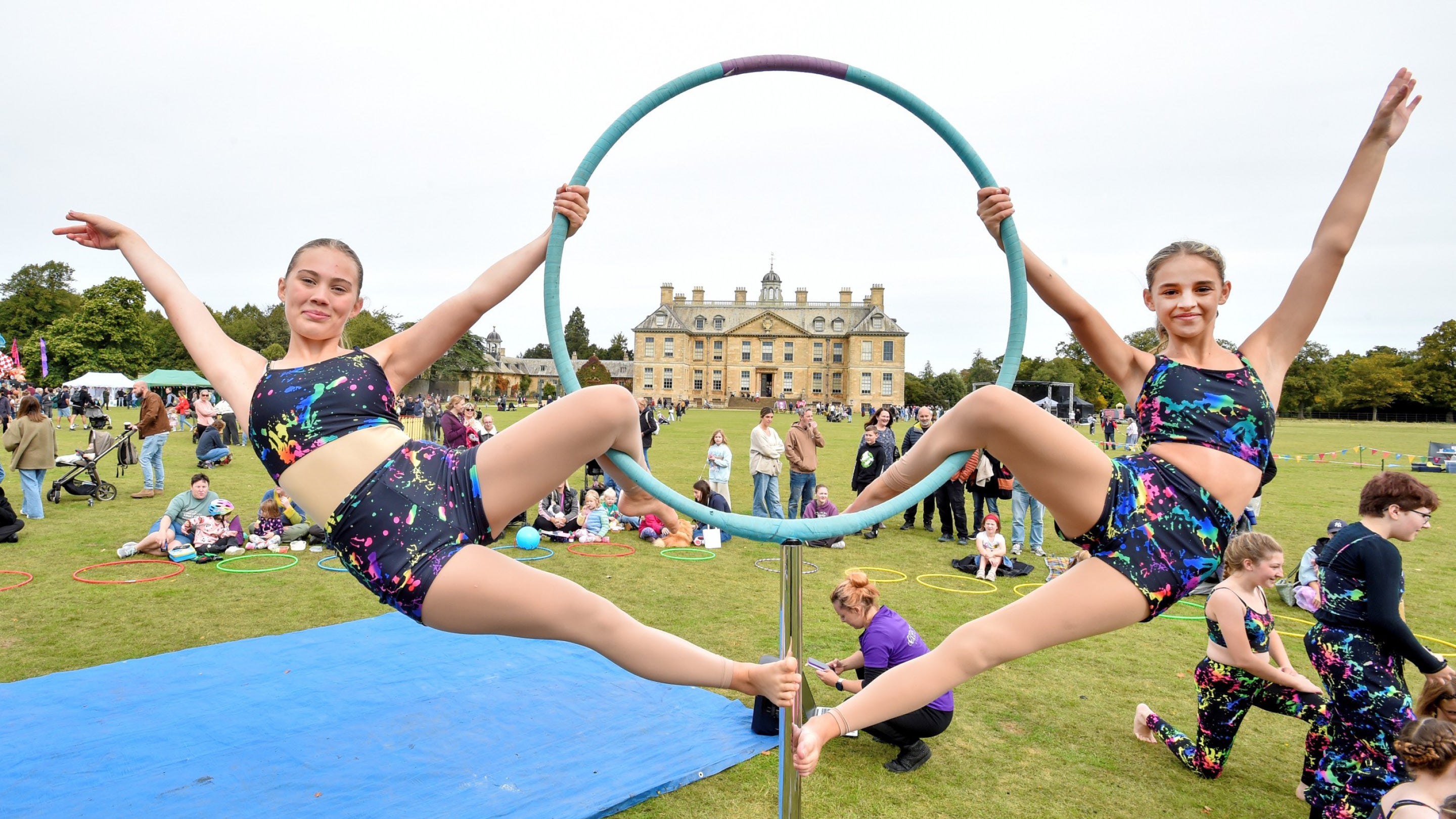 Two gymnasts hang on either side of a hoop with Belton House framed in its centre.