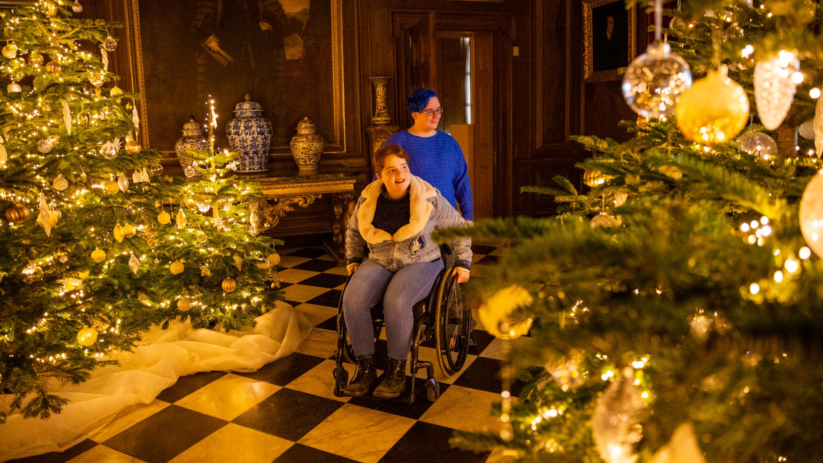 Visitors exploring festive decorations at Belton House, Lincolnshire with Christmas trees and lights in the foreground and a painting in the background