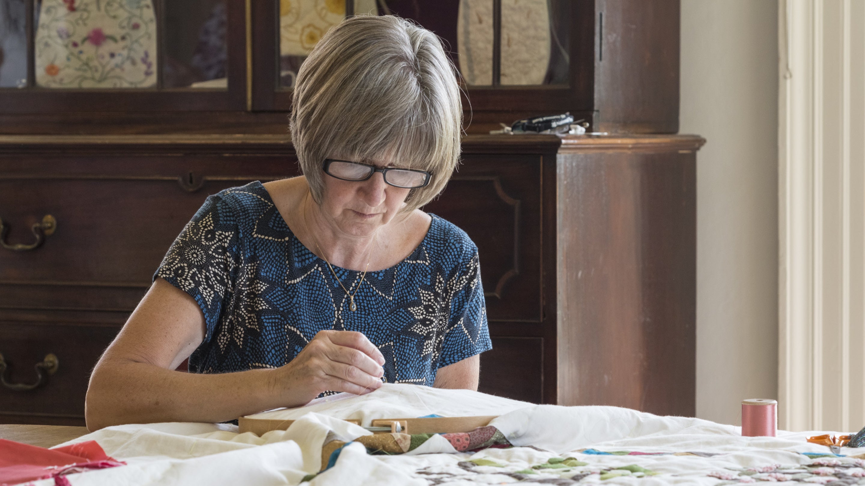 A woman with short grey hair and glasses is working on some embroidery at a table.